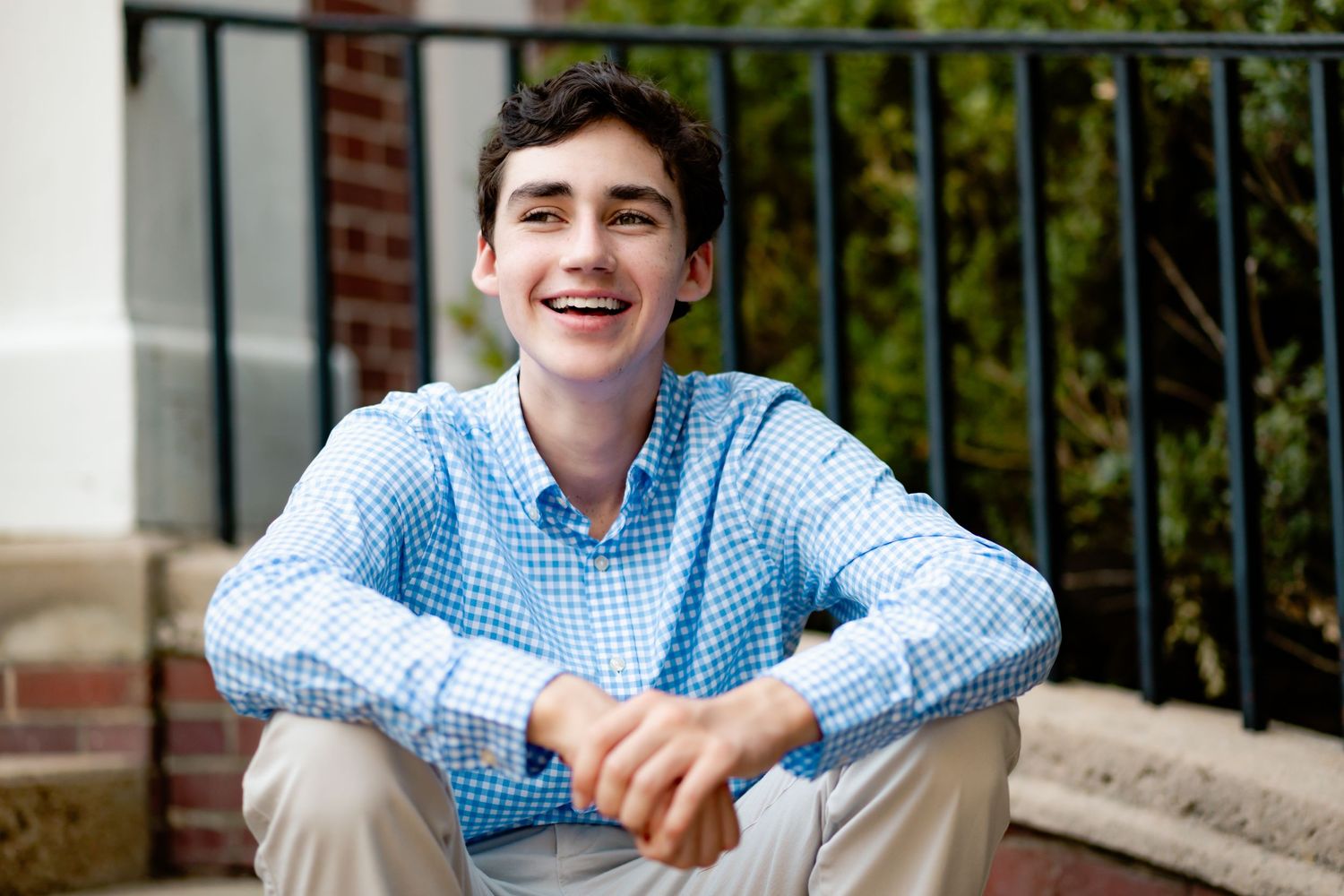 A young person in a light blue checkered button-down shirt sits smiling outdoors by a metal railing.