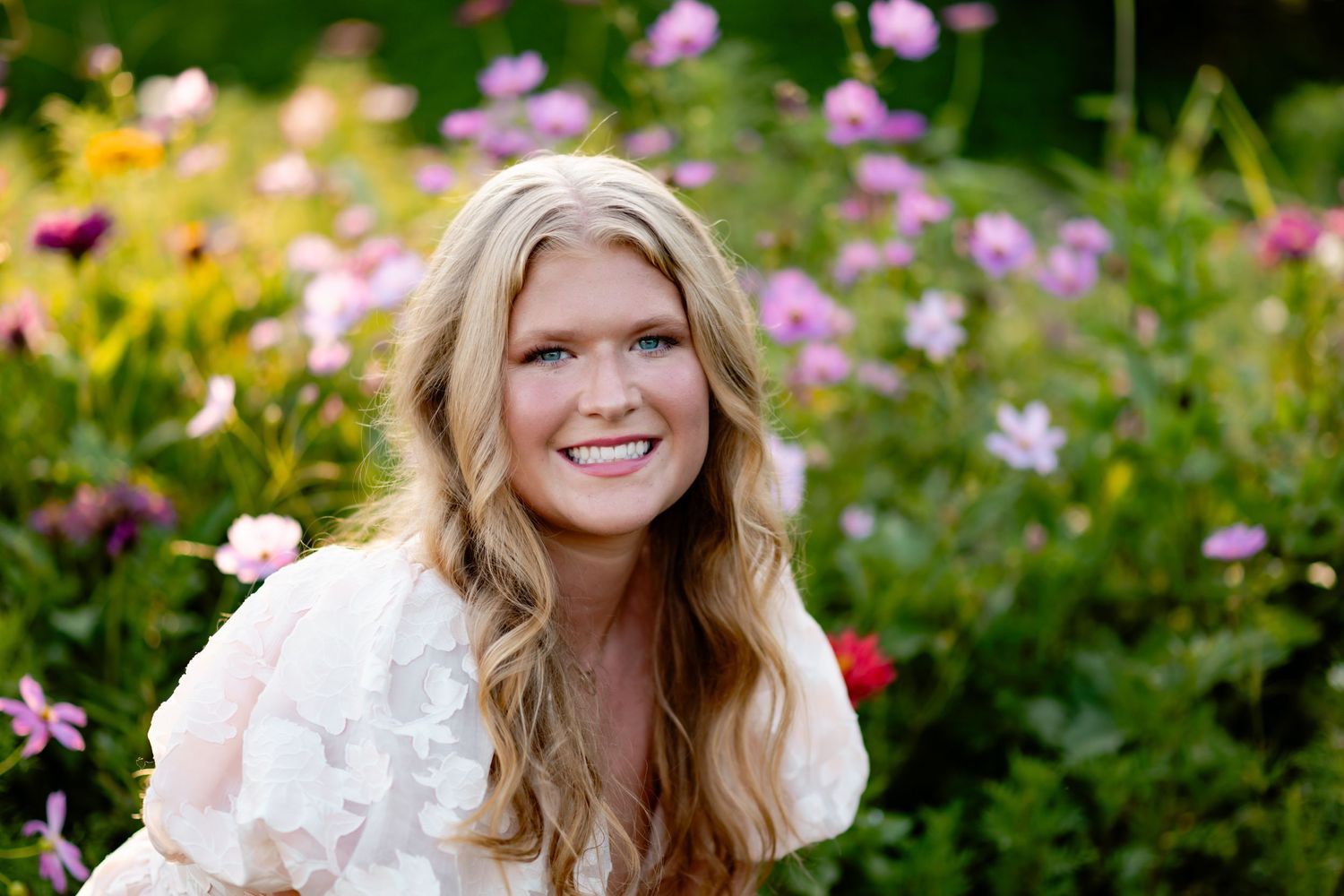 Person wearing white lace dress smiles joyfully while posing among blooming pink and yellow flowers in garden setting.