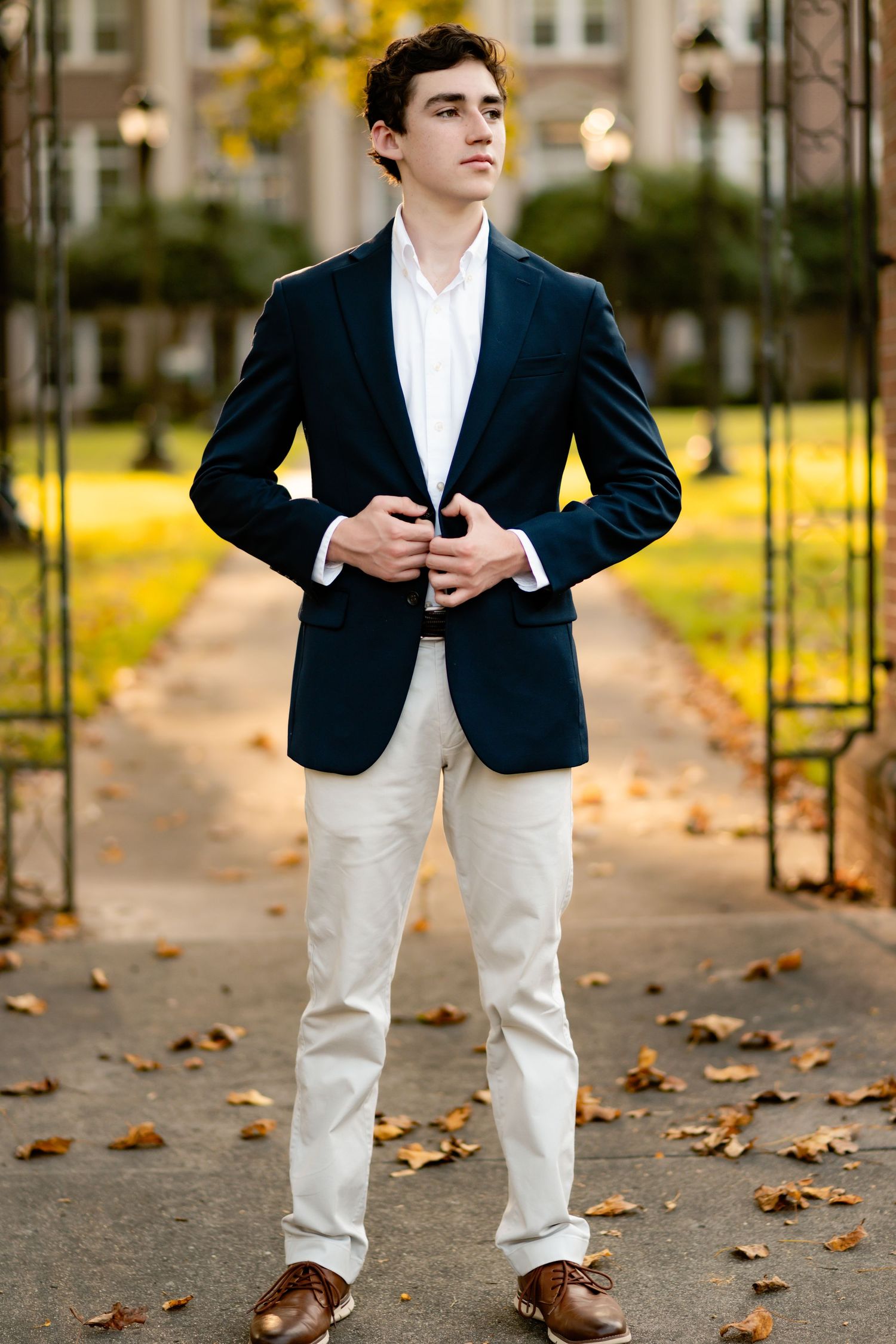 A person in a navy blazer and khaki pants stands on a leaf-covered path during autumn at golden hour.