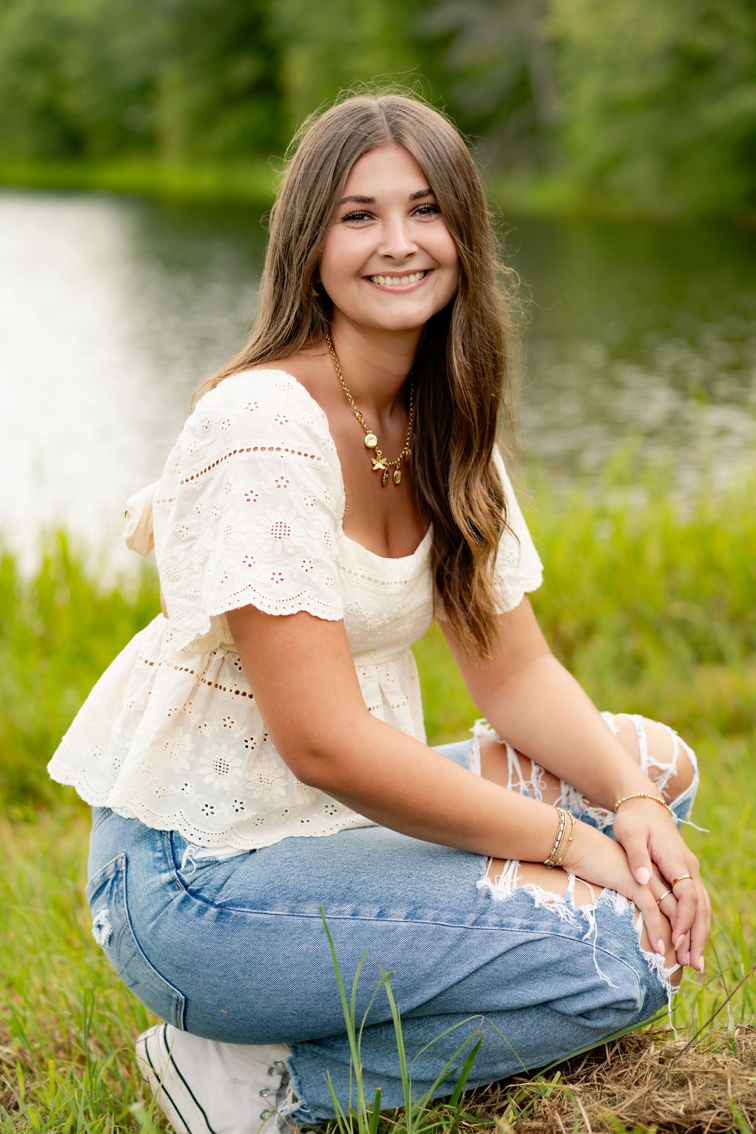 Someone in white eyelet top and jeans sitting by pond on summer day.