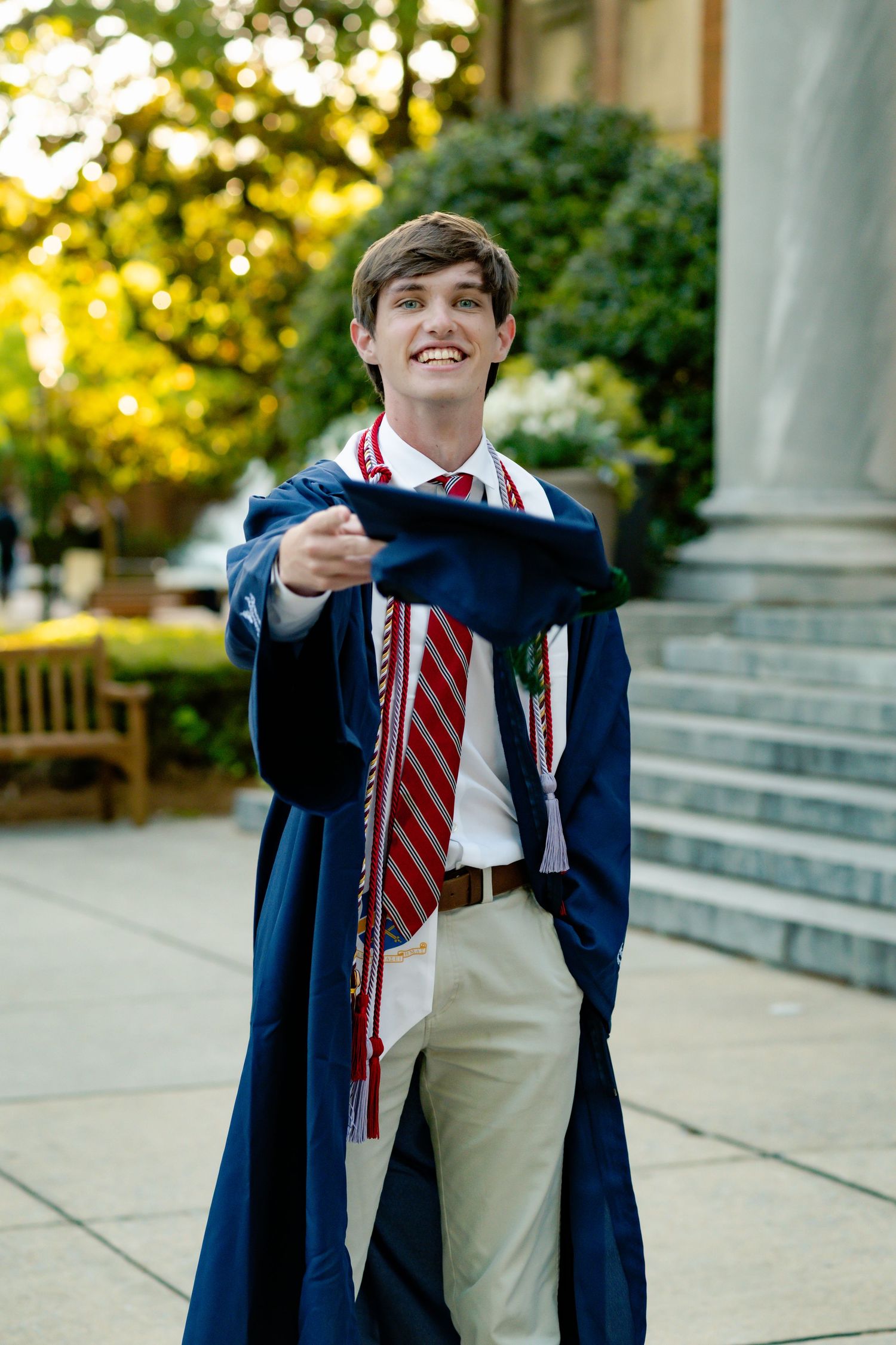 Graduate in navy cap and gown poses with his diploma on university steps at sunset.