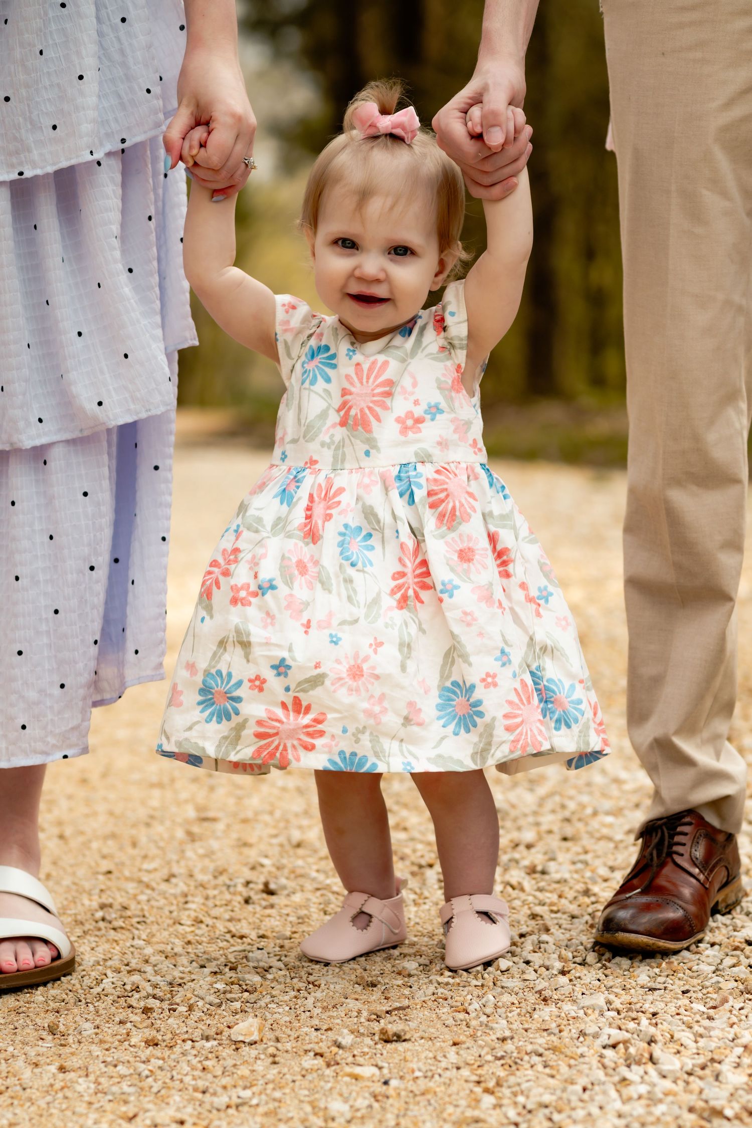 Small child in floral dress holding parents' hands while learning to walk on path.