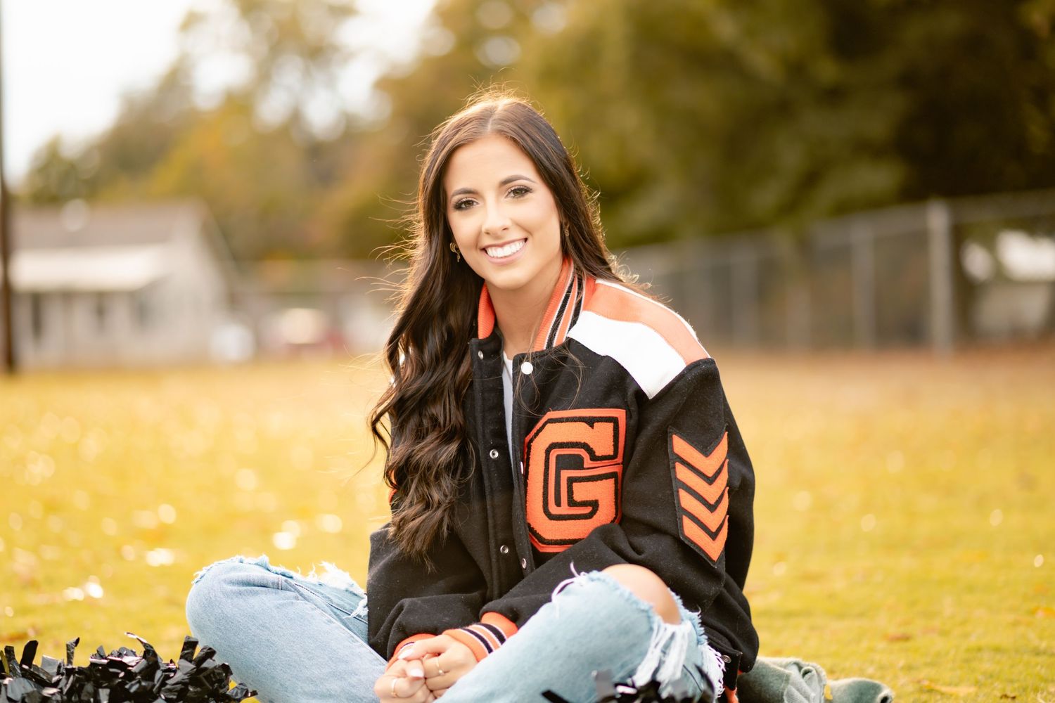 A cheerleader in a varsity letterman jacket sits on grass during a sunny autumn afternoon.