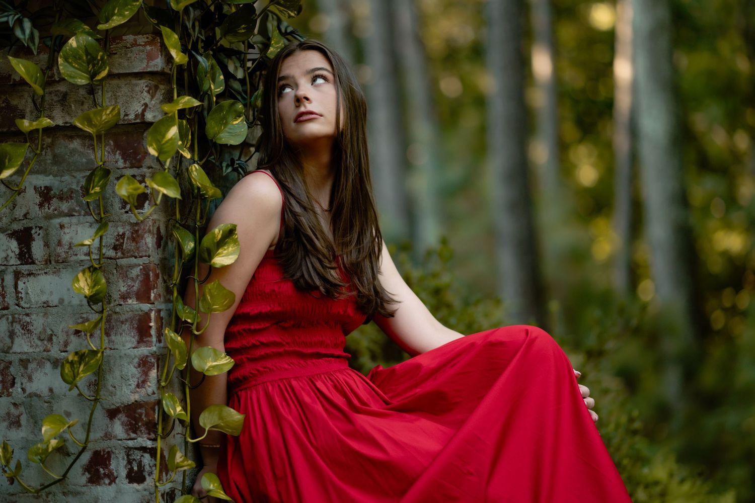 Figure in flowing red dress poses thoughtfully against stone wall in lush forest setting.