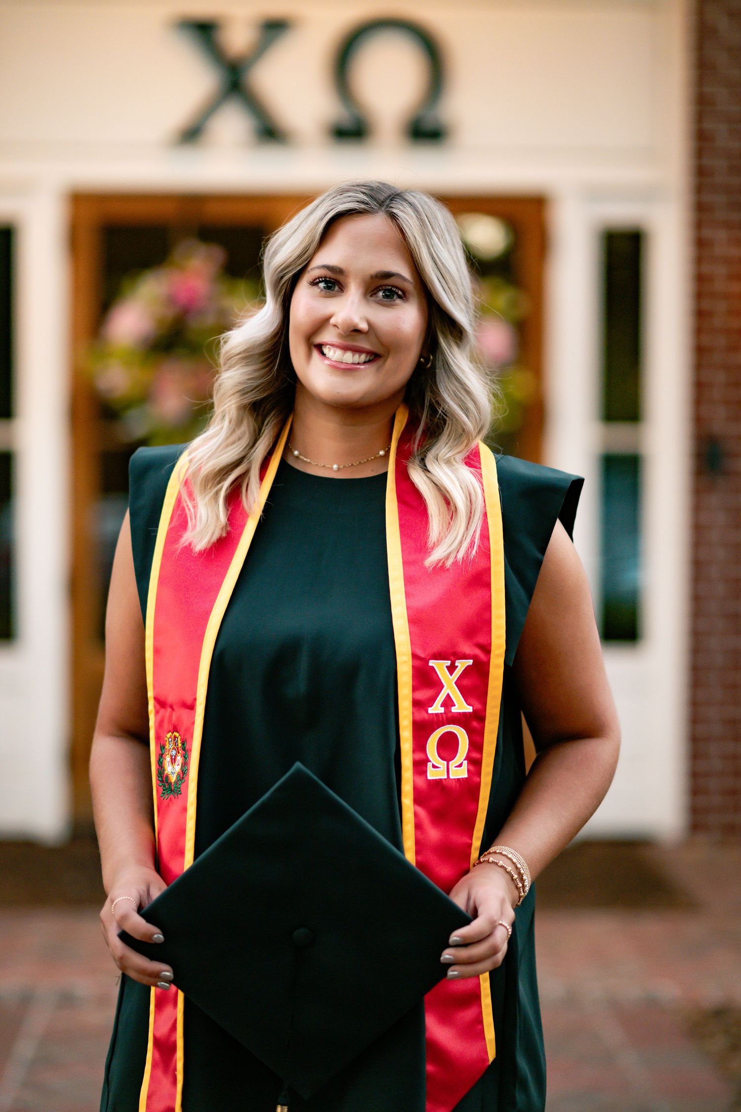Graduate wearing black dress and red honor stoles smiles during outdoor photoshoot.