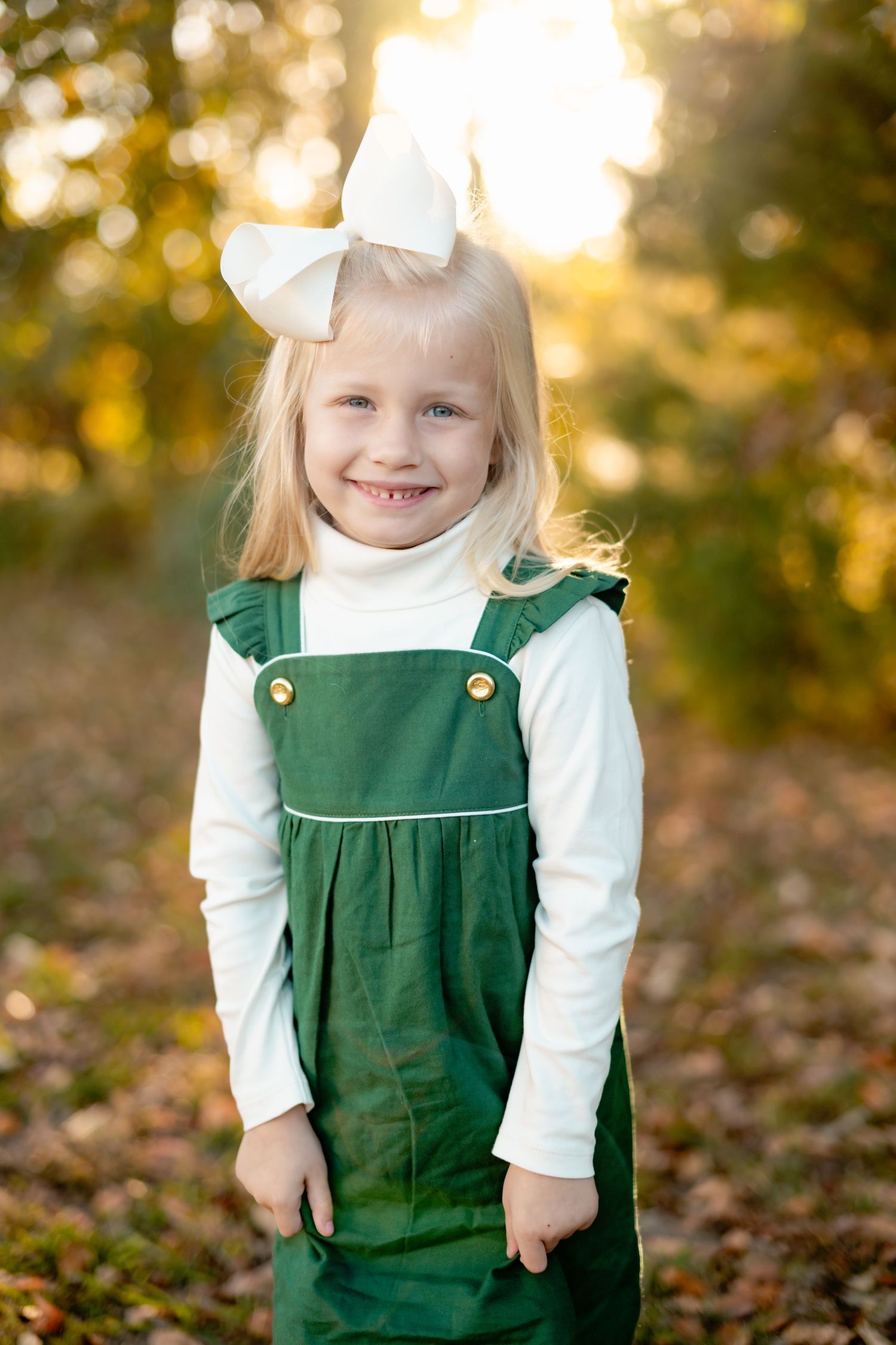 Child wearing a green corduroy jumper dress with white bow and blouse against autumn foliage background in sunlight.
