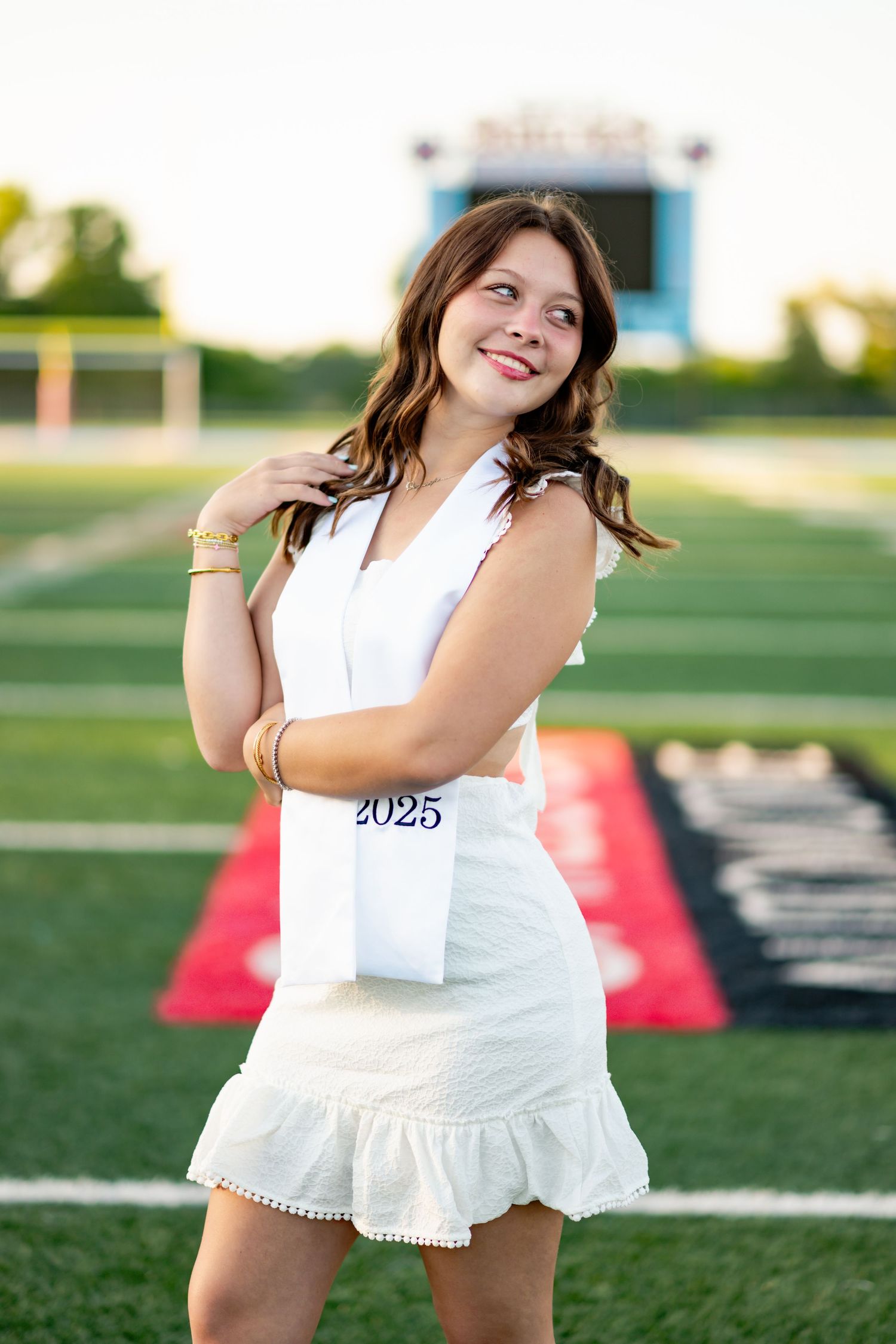 Graduate wearing white dress and sash poses playfully on outdoor field during sunset celebration.