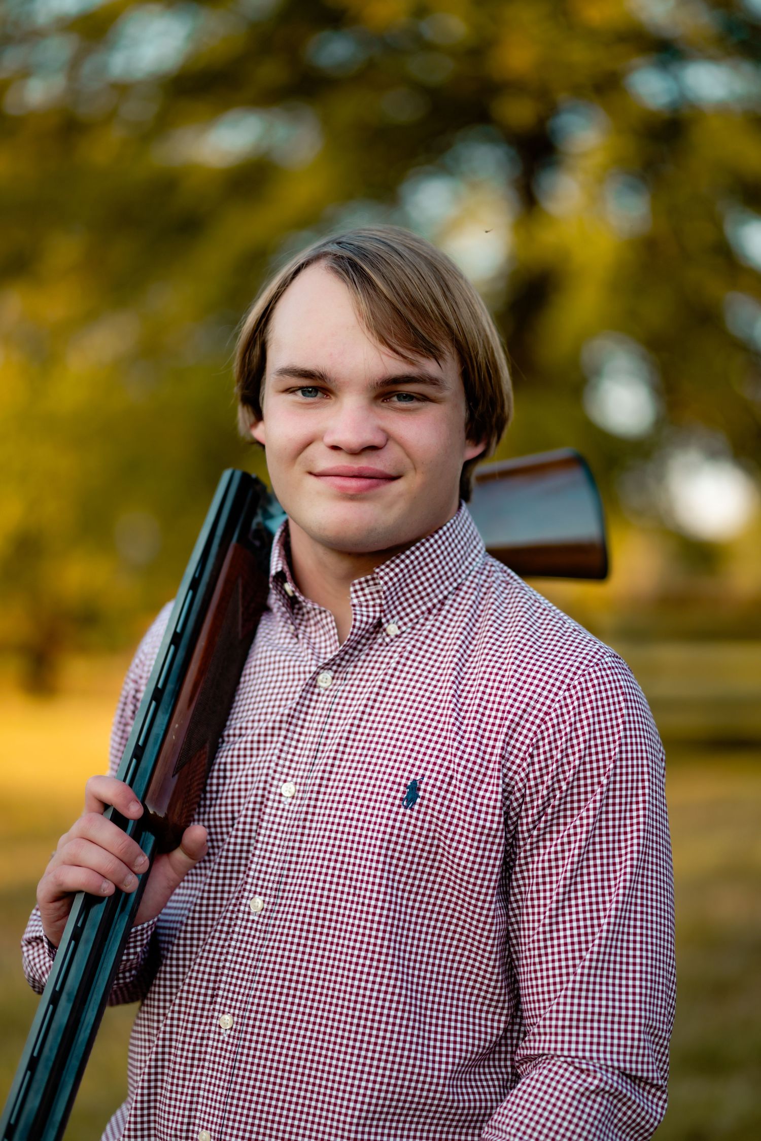 Person in red and white checkered button-down shirt poses outdoors with hunting rifle in golden afternoon light.