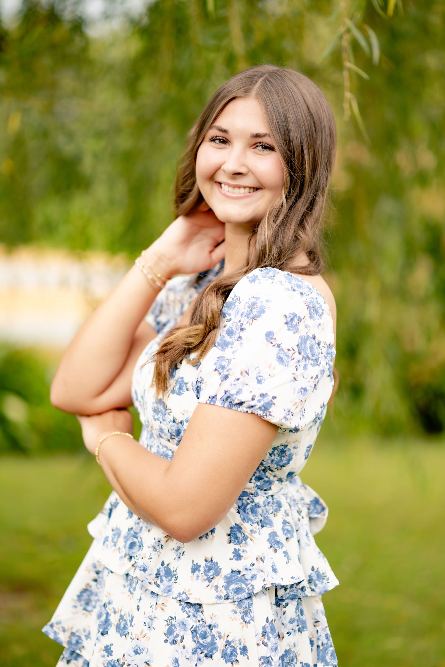 Portrait of someone in blue and white floral dress posing outdoors with willow trees.