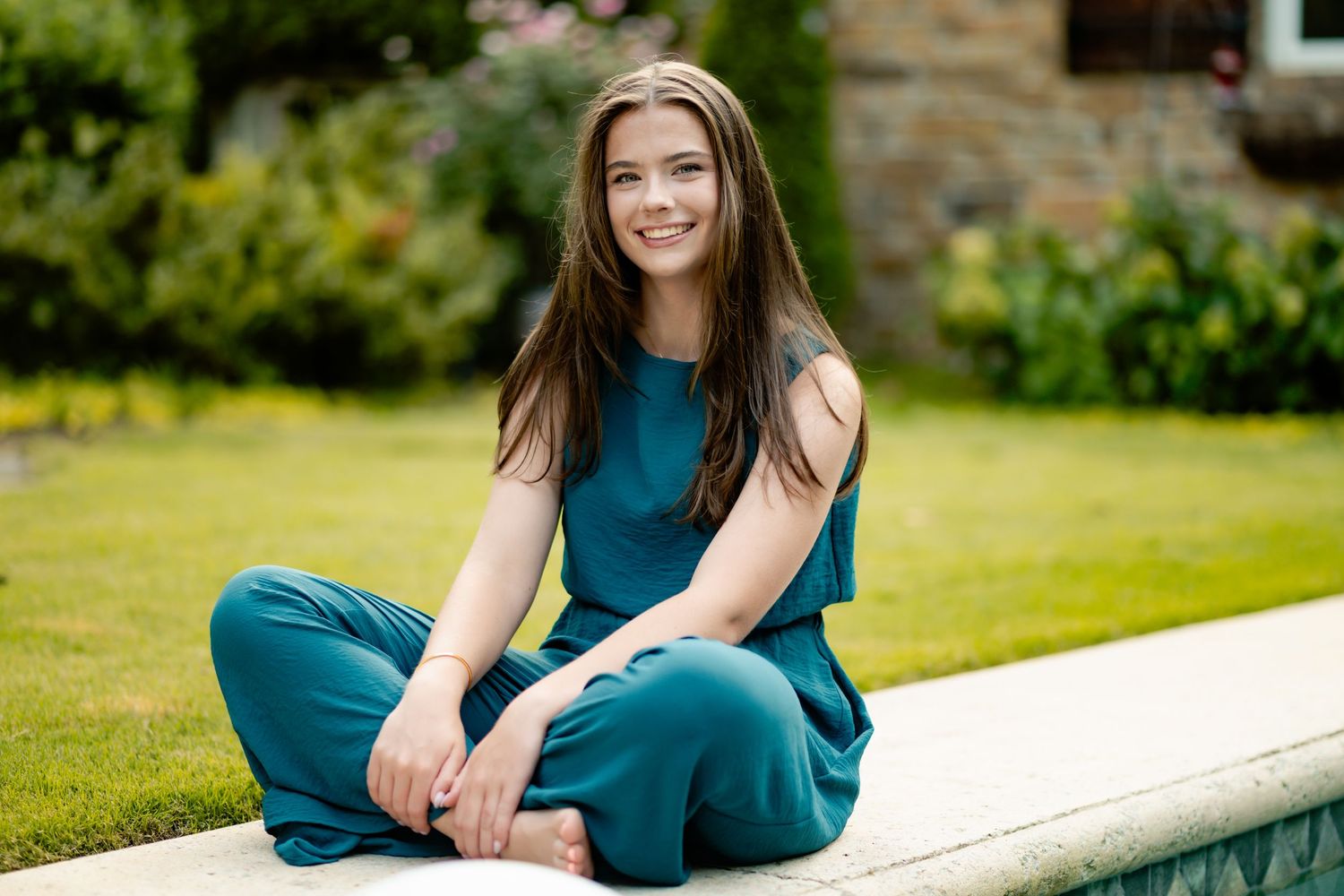 A woman in a dark teal dress sits outdoors on a white railing surrounded by a lush green garden setting.