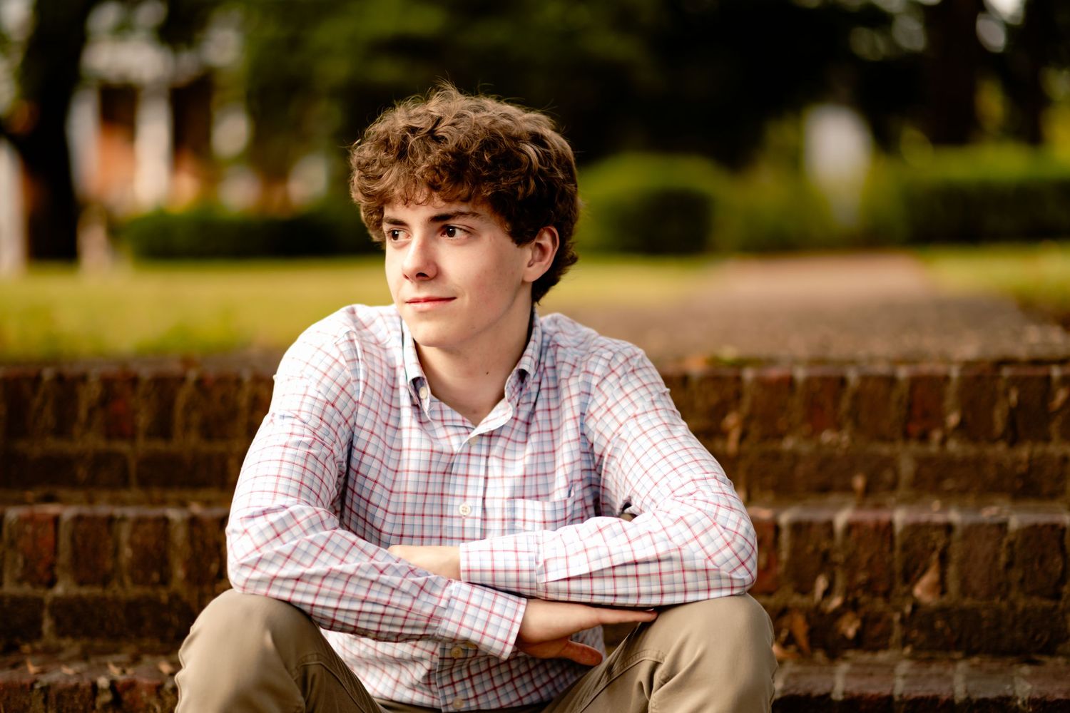 Person in a checkered shirt sits on outdoor brick steps against a natural backdrop of trees and greenery.