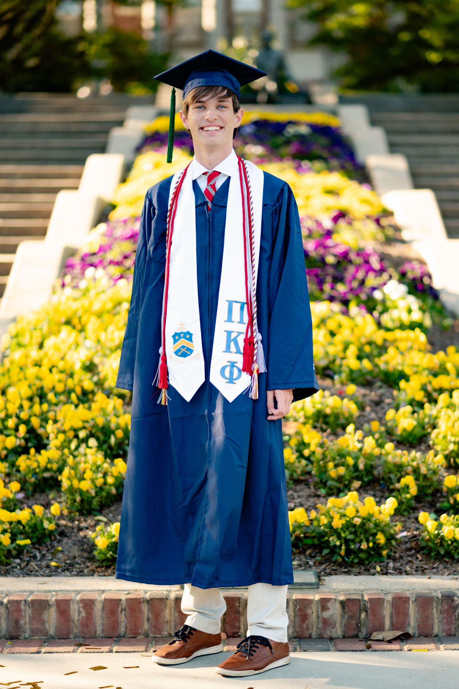 Graduate in navy cap and gown with decorated stoles poses by yellow flowers.