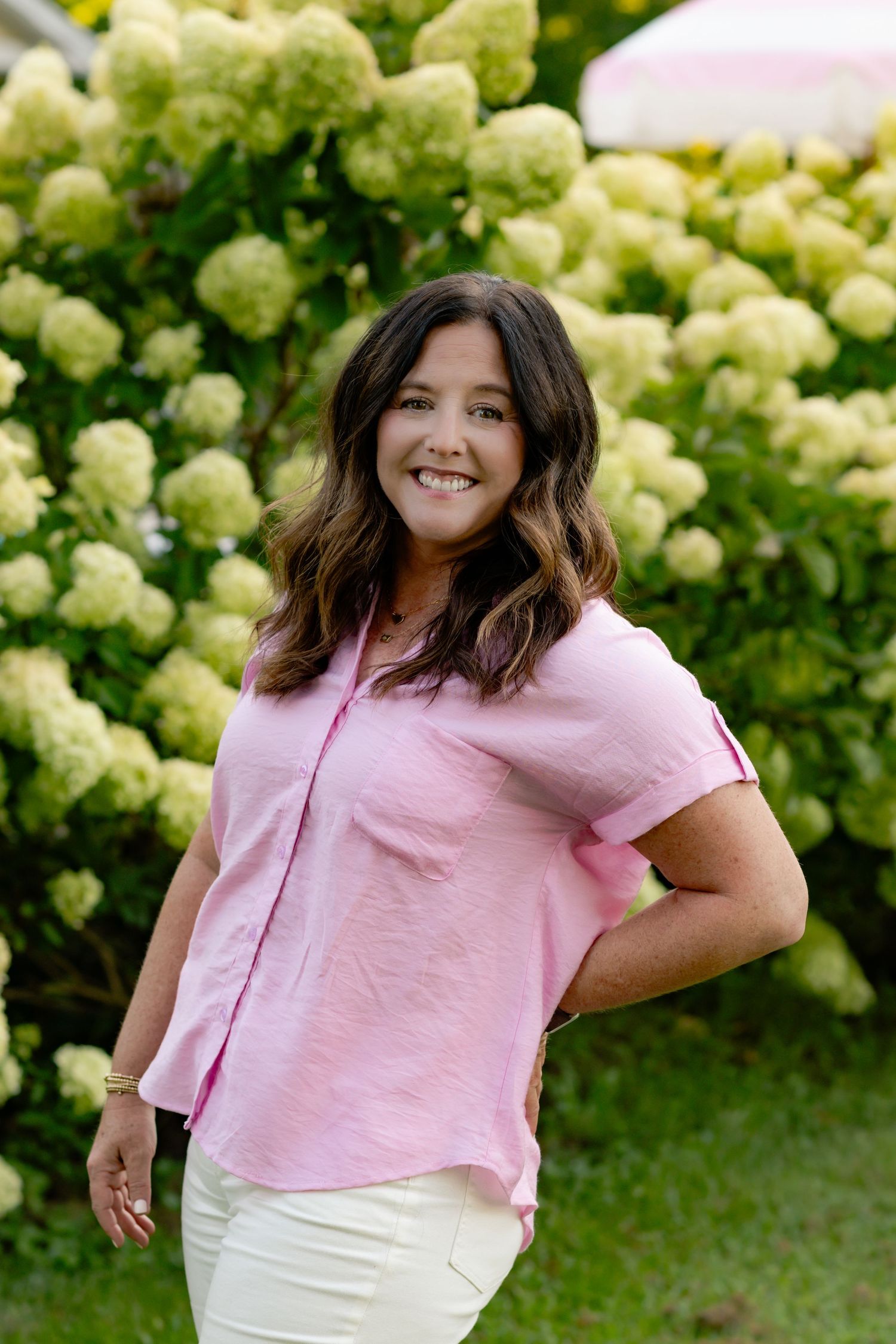 A person in a pink t-shirt poses playfully in front of lush white hydrangea bushes on a summer day.