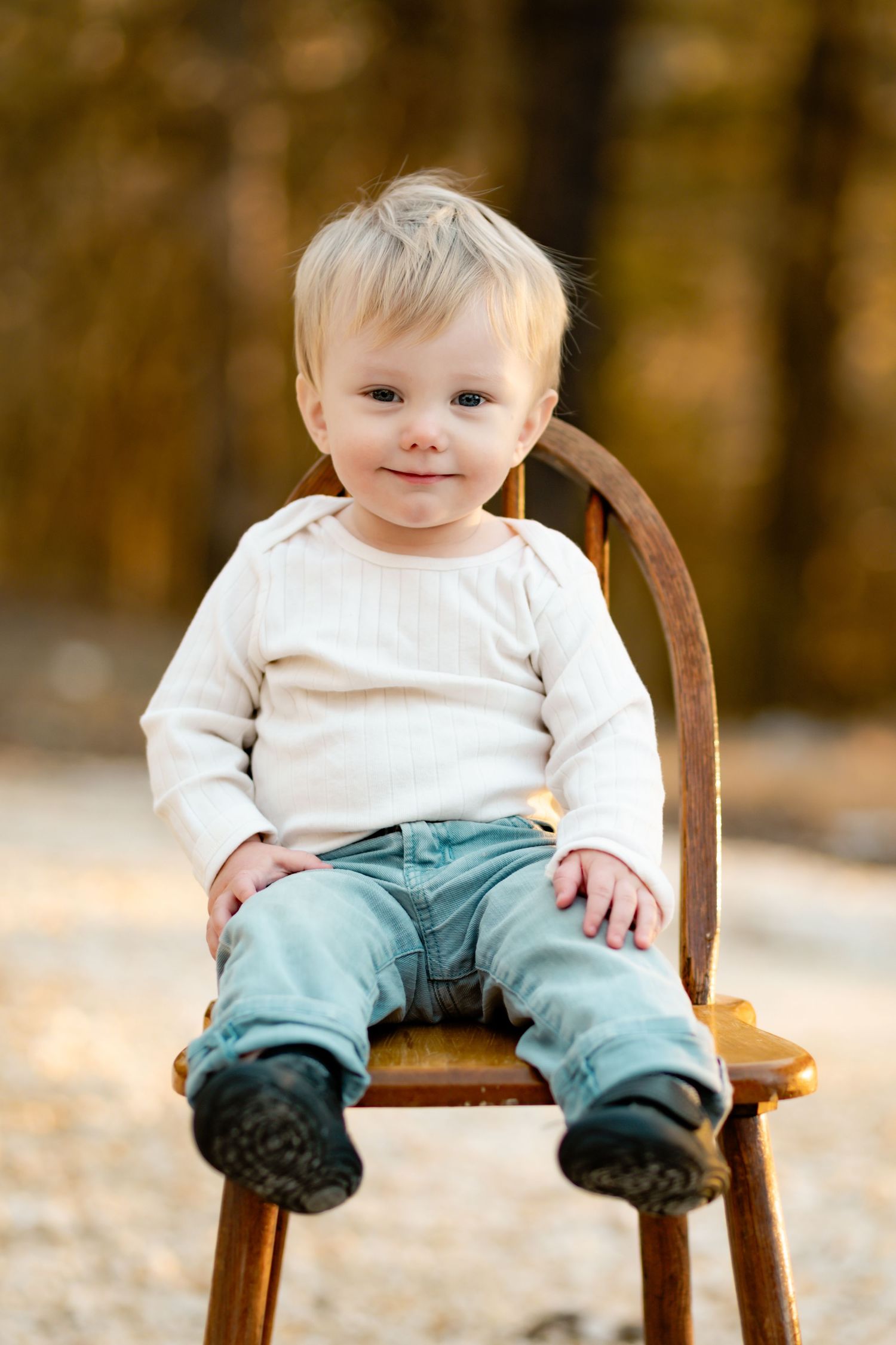 Small child wearing white sweater and blue pants sitting on wooden chair in autumn setting.