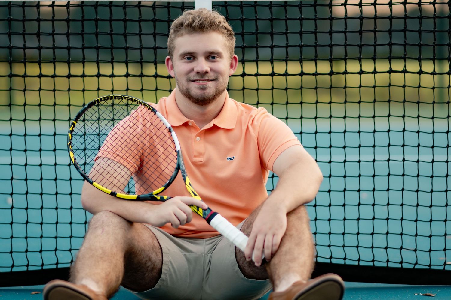 A tennis player in an orange shirt stands behind a green net on a tennis court during a match.
