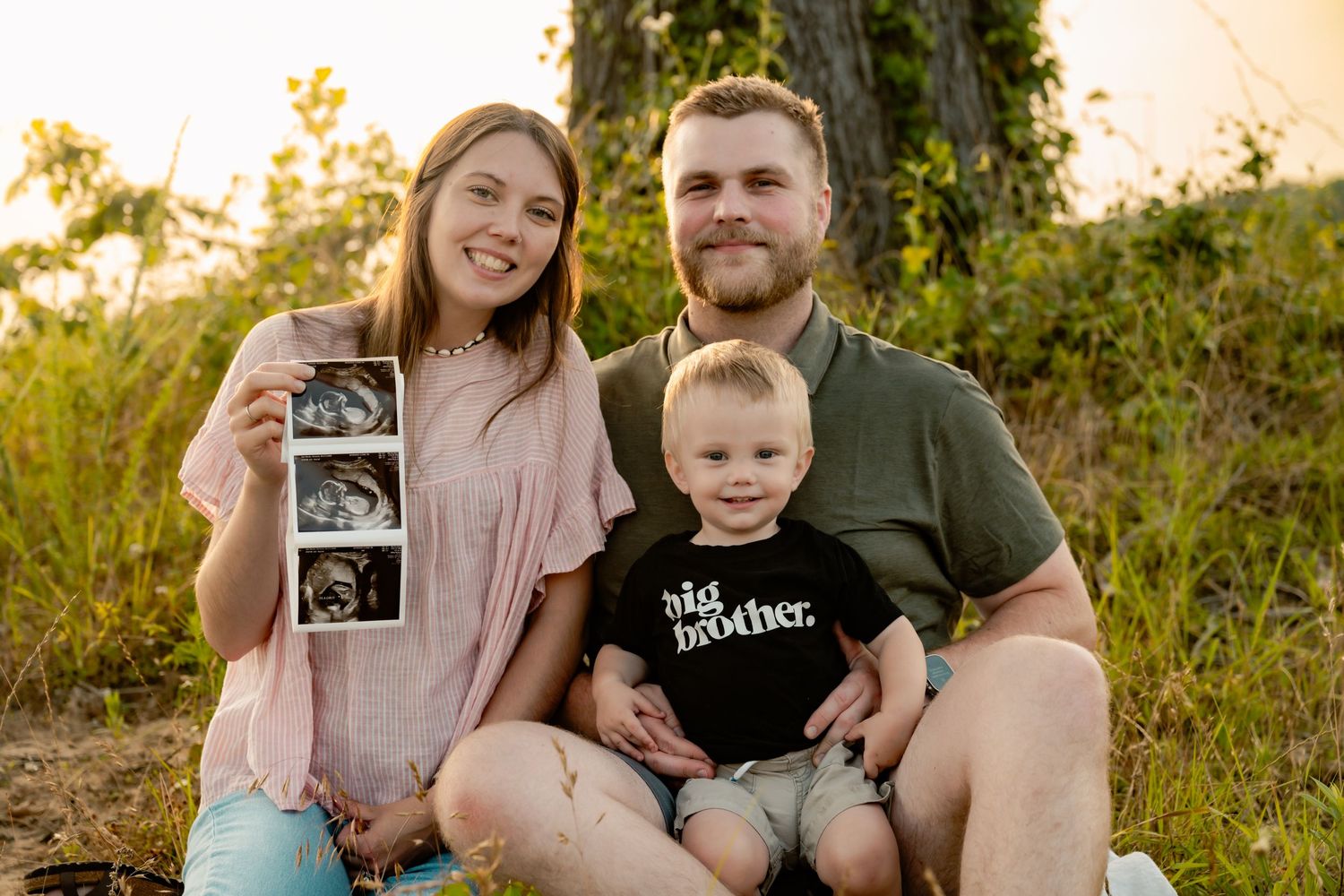 Family sitting outdoors holding ultrasound photos while announcing pregnancy.