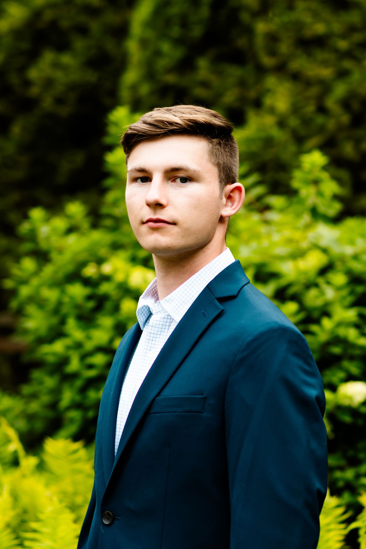 Professional headshot portrait of person in navy suit against green natural outdoor background.