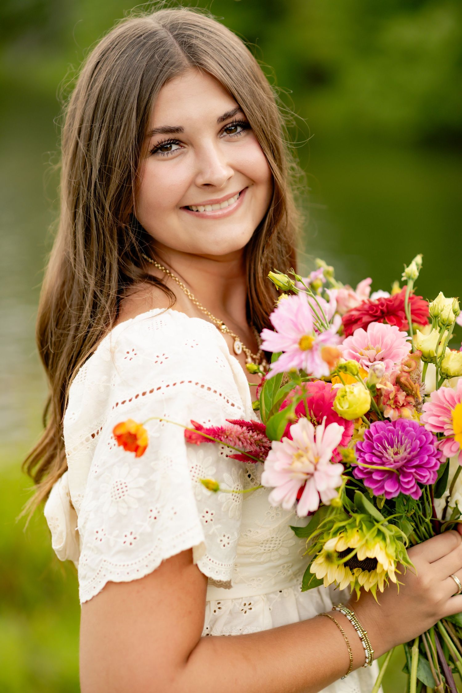 Person wearing white eyelet dress holding vibrant summer bouquet with pink, red and yellow flowers in outdoor setting.
