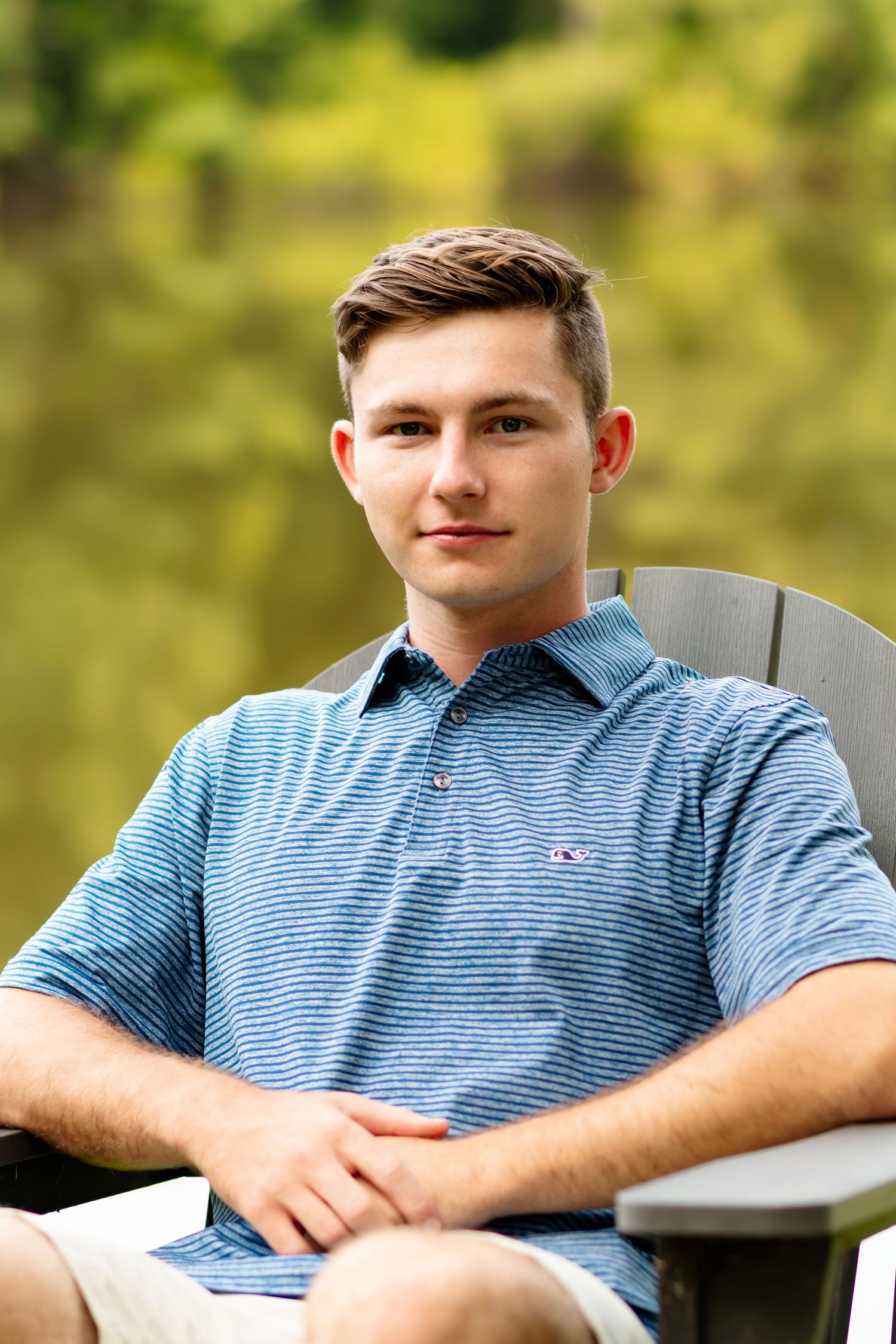 Young man in blue striped polo shirt sits outdoors in a casual pose against natural blurred background.