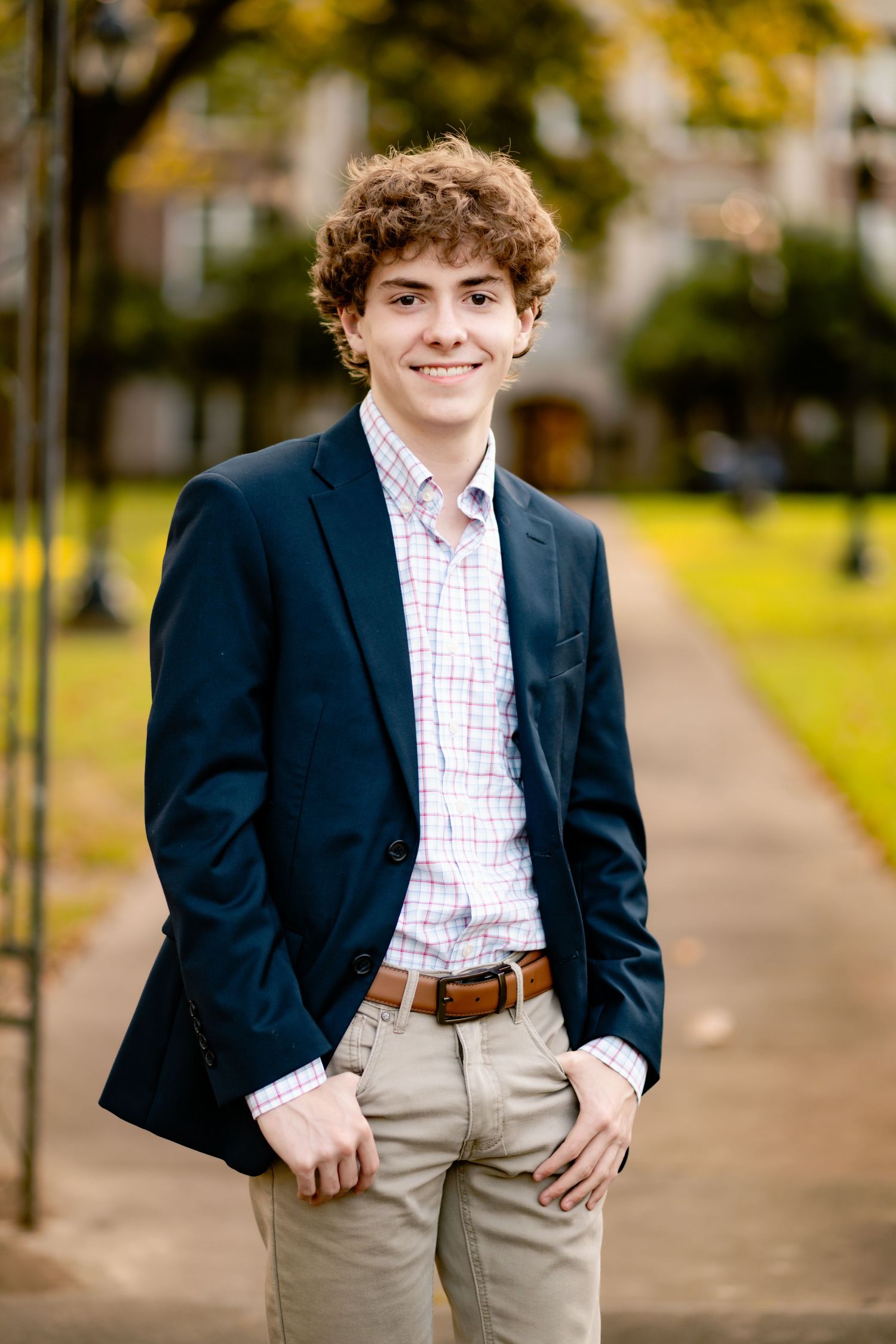 A young student in a navy blazer and khaki pants poses casually on a tree-lined walkway during an autumn photoshoot.