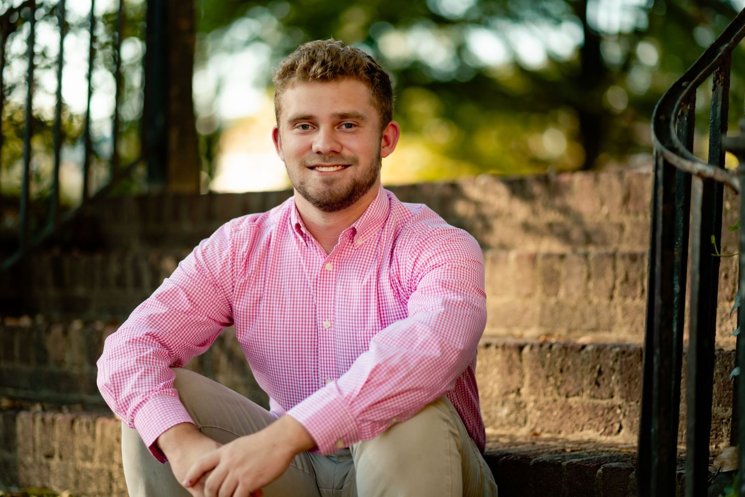 Young person in pink button-down shirt sitting outdoors with blurred foliage and fence in background.