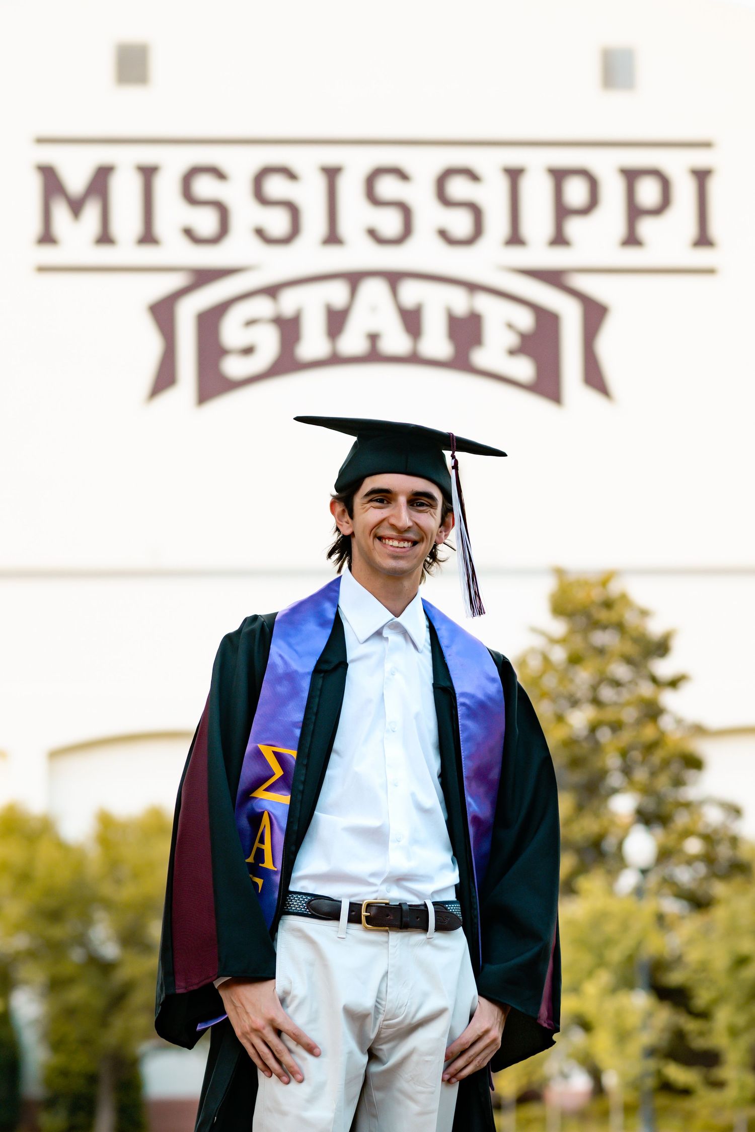 A graduate wearing blue and purple regalia stands smiling in front of a Mississippi State University sign.