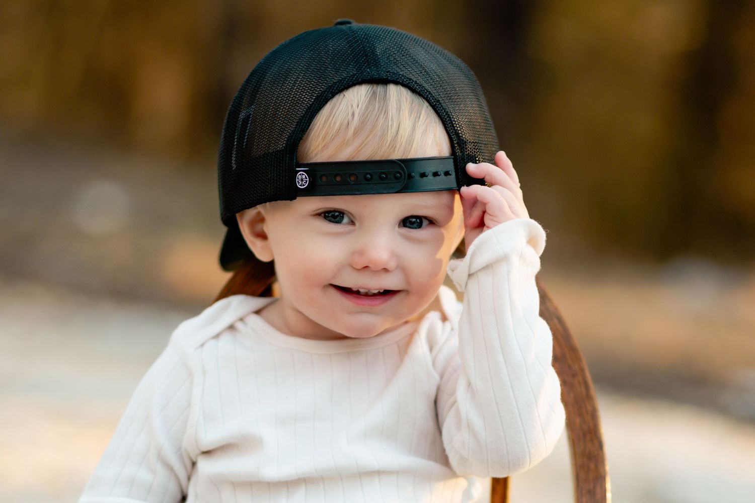 Small child in white top wearing backwards baseball cap while smiling.