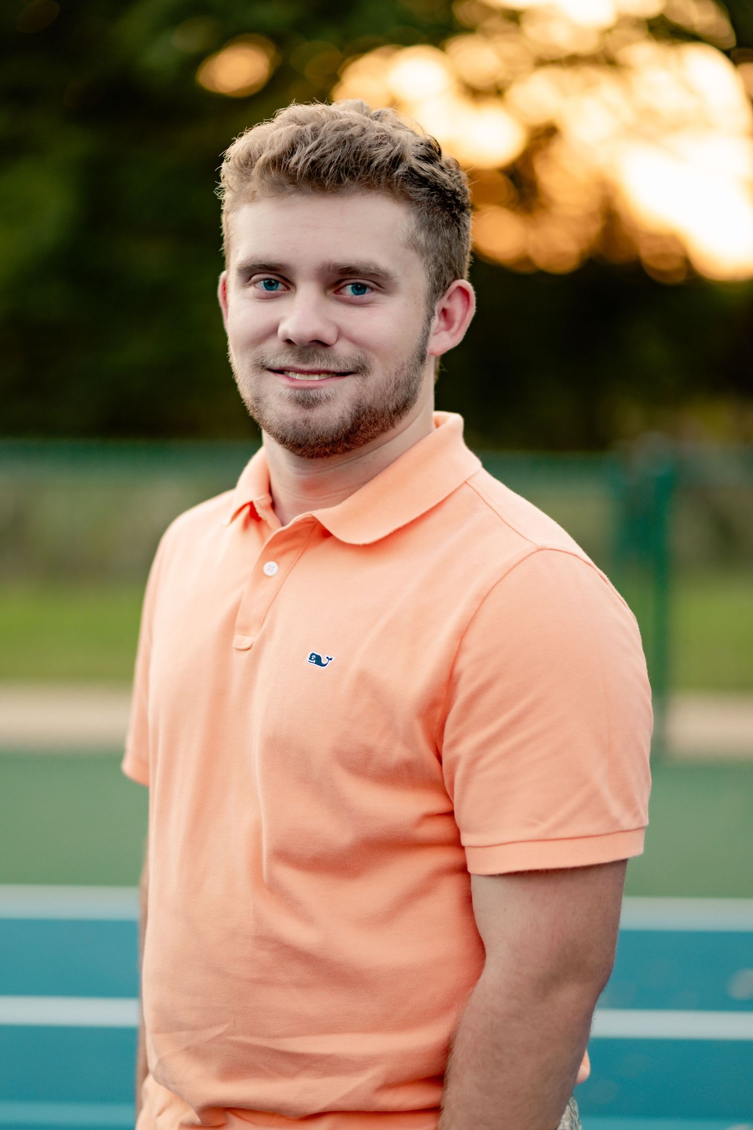 Man wearing peach-colored polo shirt smiles while standing outdoors against blue surface at sunset.
