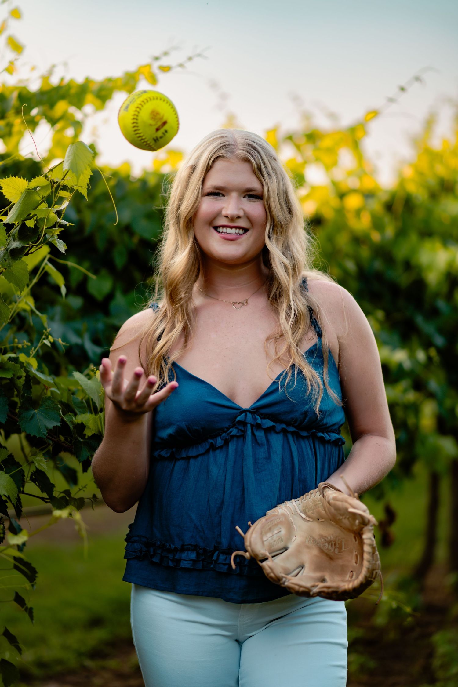 A woman in a blue top poses playfully with a softball in a sunny vineyard setting.