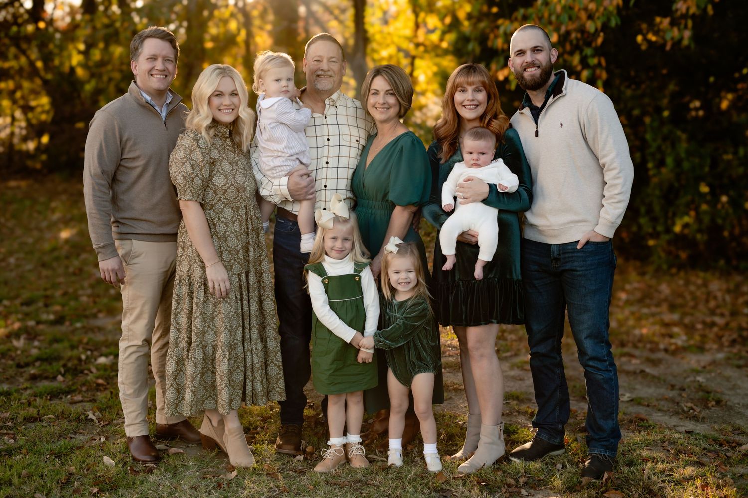 A family group poses outdoors in coordinated green and neutral clothing against a warm autumn backdrop with golden foliage.