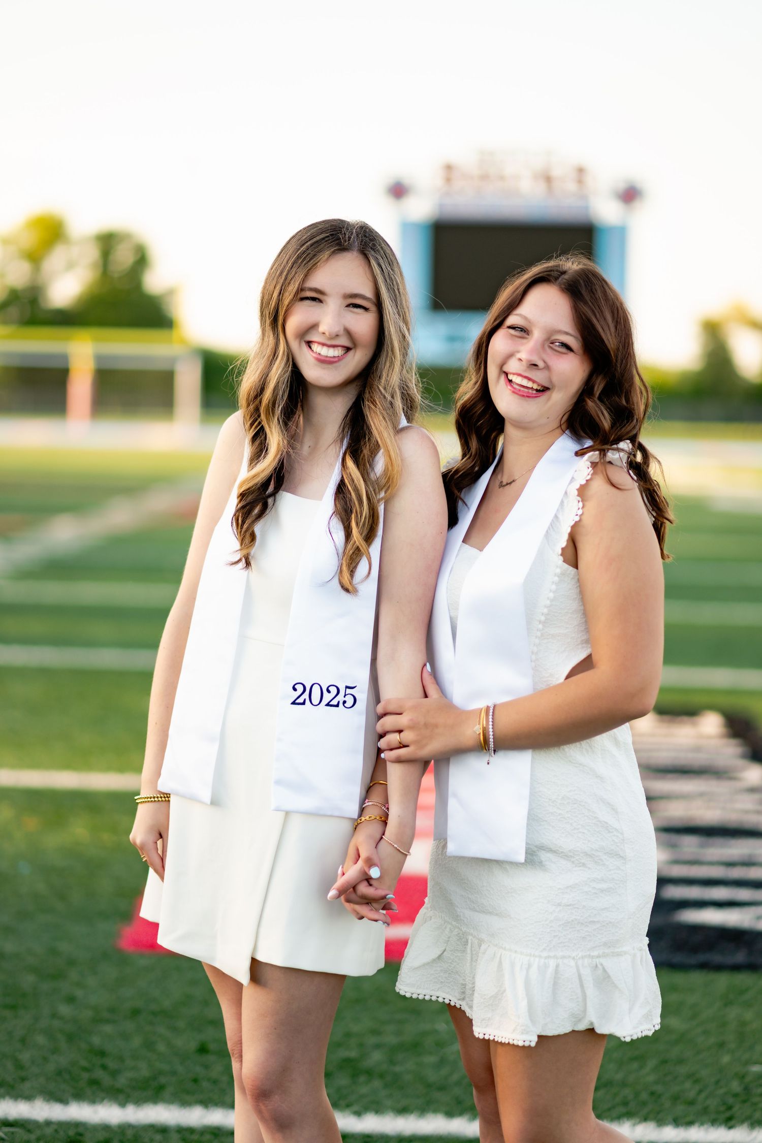Two people in white summer outfits pose together on an outdoor sports field.