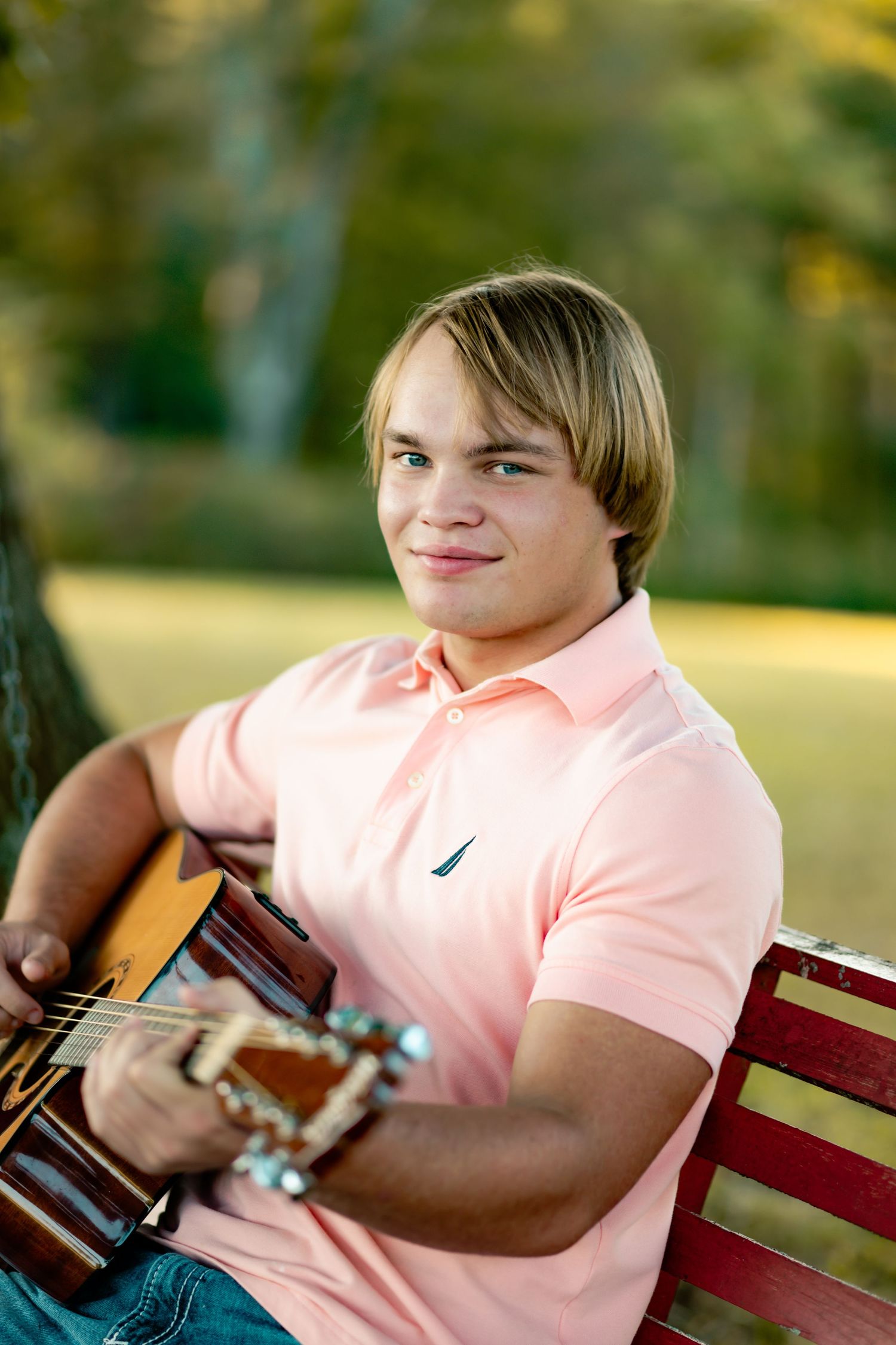 A person wearing a pink polo shirt sits on a red bench while playing an acoustic guitar outdoors.