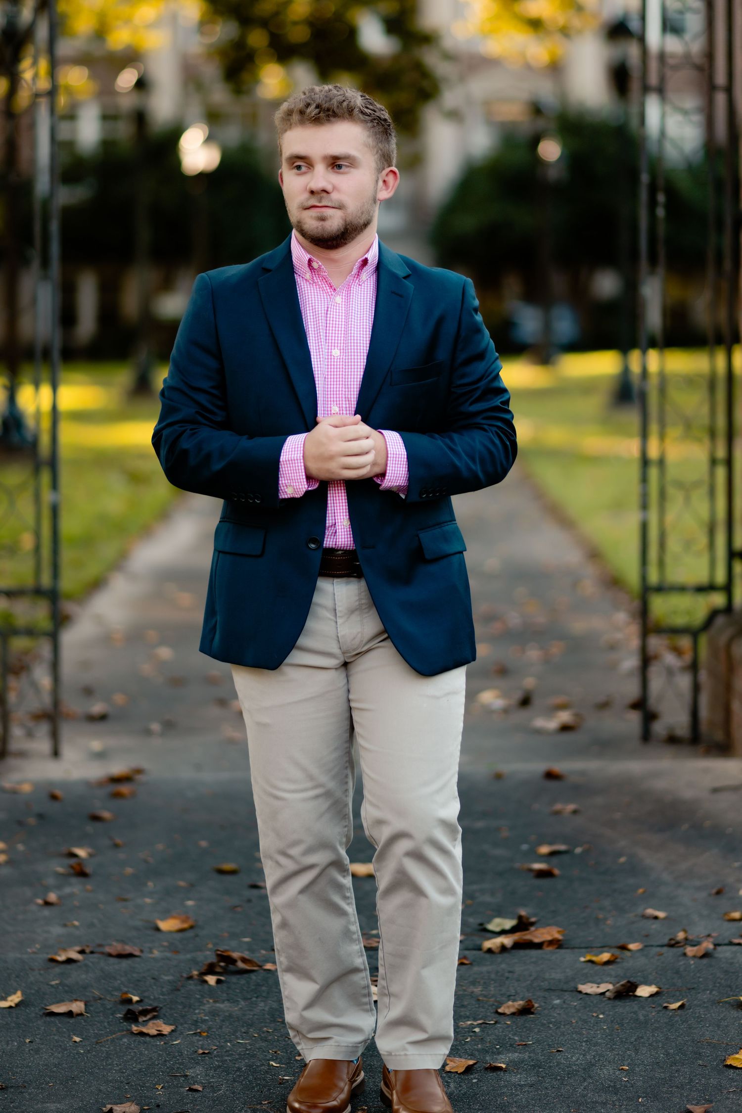 Man in navy blazer and pink dress shirt poses casually on leaf-covered path during autumn.