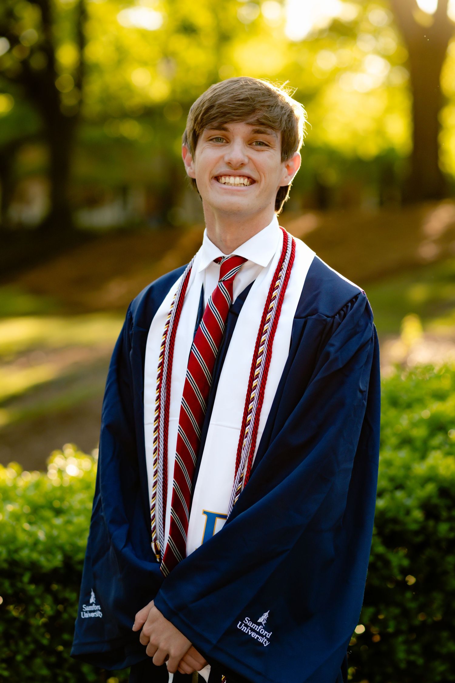 Graduate in navy cap and gown with striped stole poses outdoors.