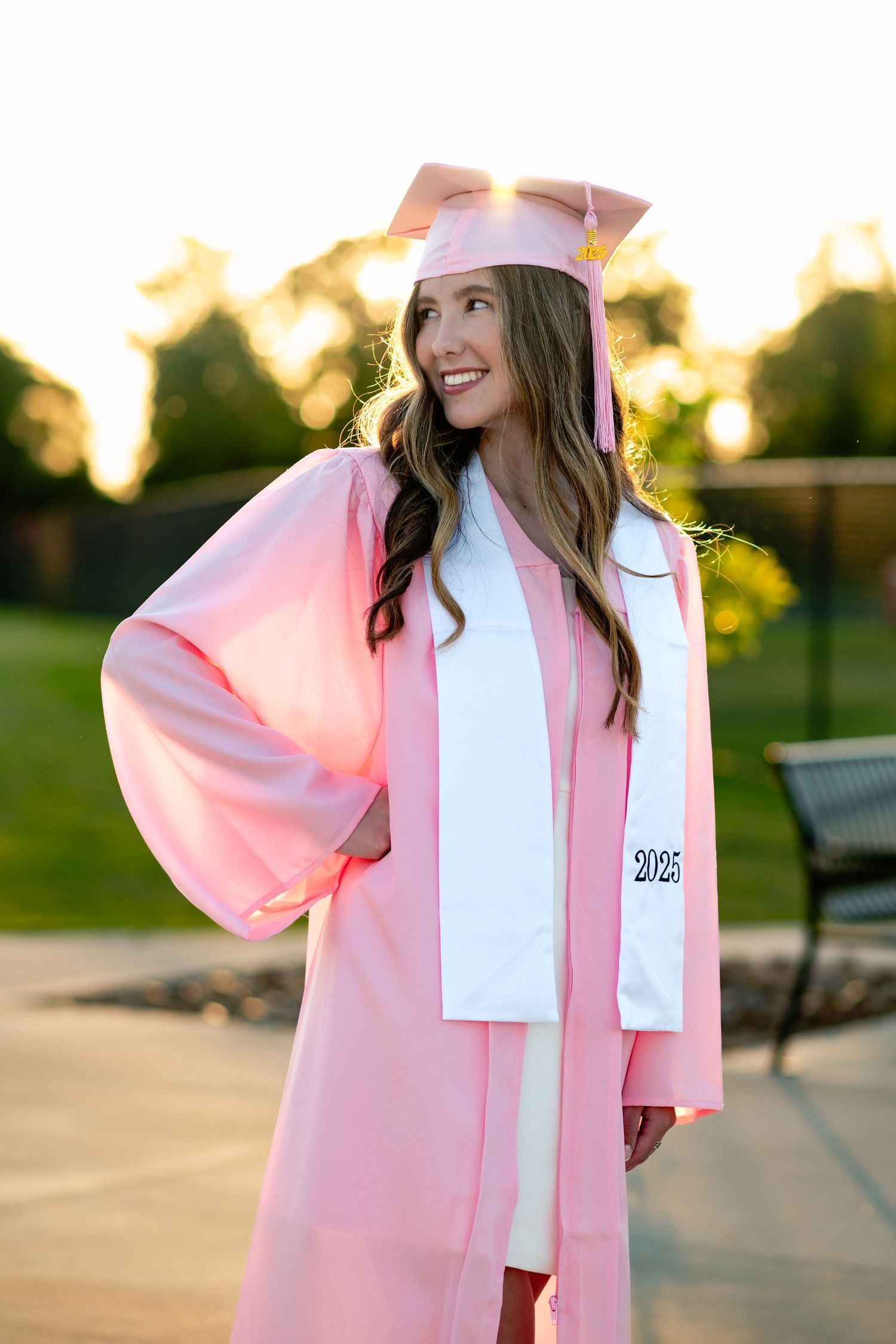 Graduate in pink gown and white sash smiles during golden hour photo session.
