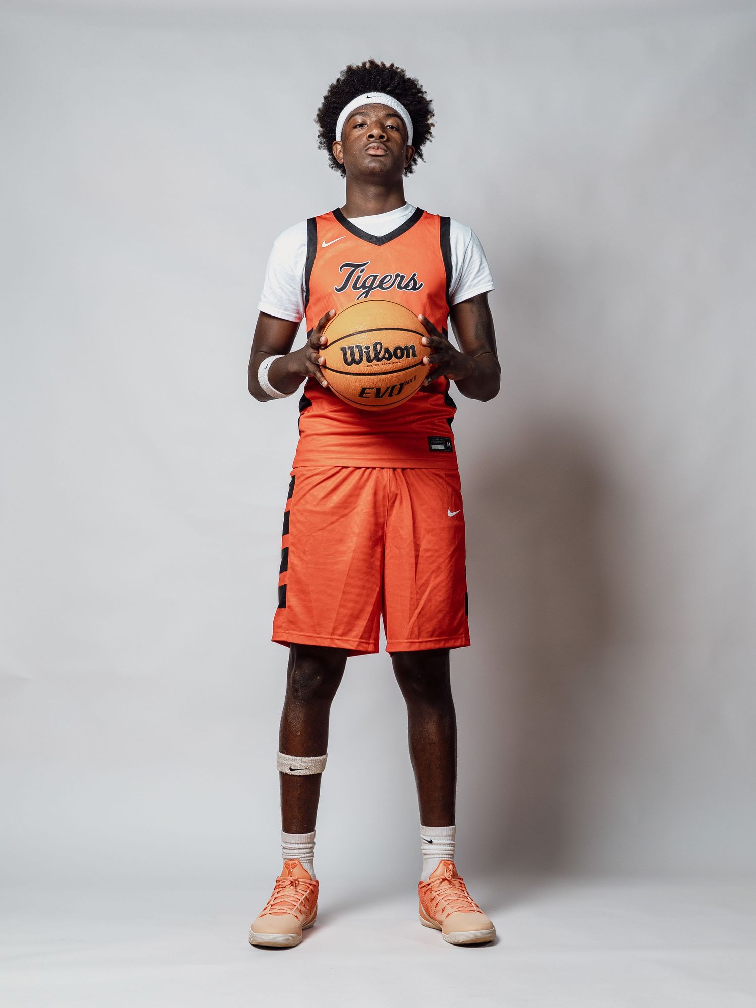 Basketball player in an orange Tigers jersey and shorts, white headband, holding a Wilson basketball against a plain gray studio background.