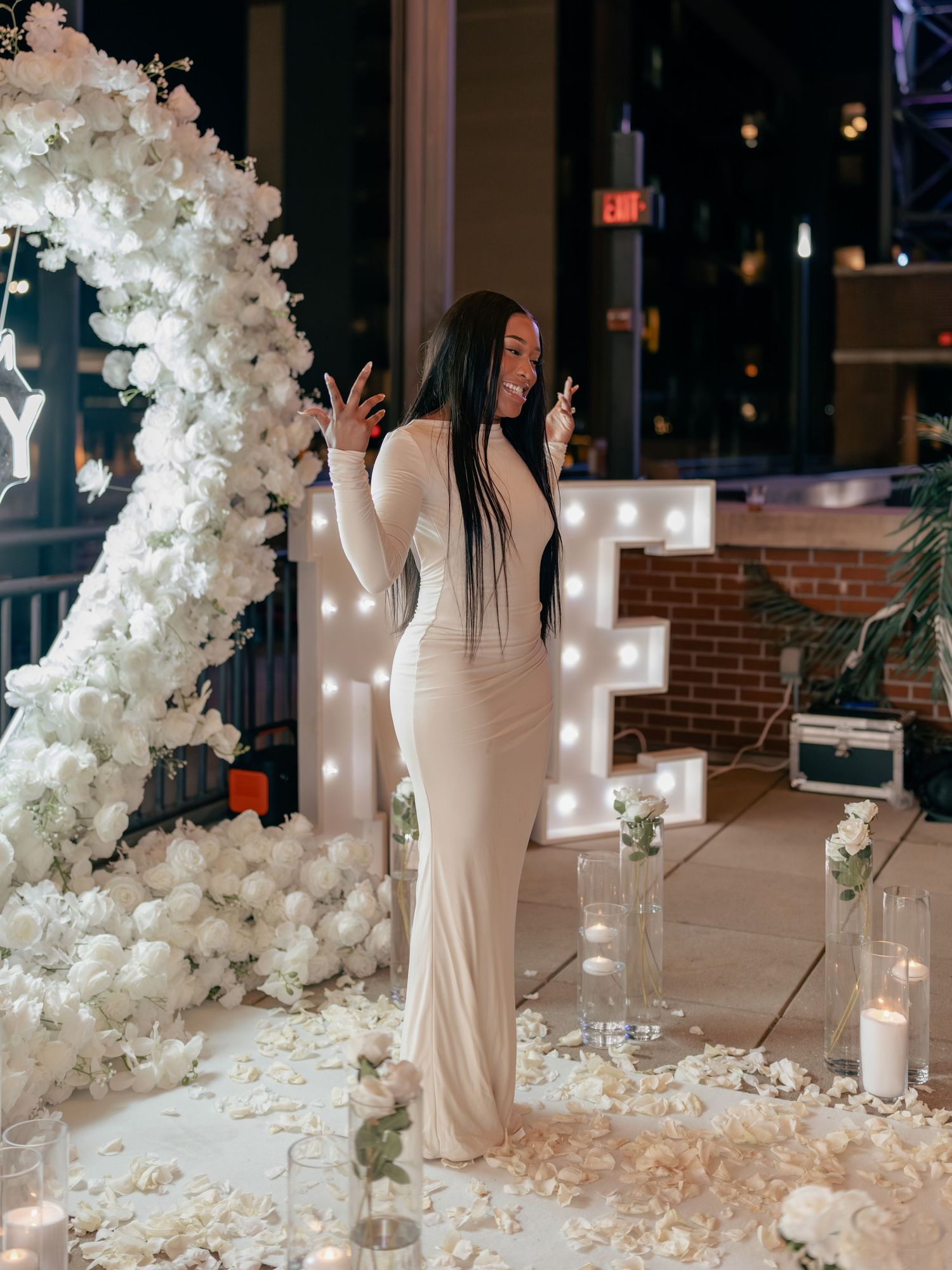 Woman in elegant cream gown poses by white floral arch with candles and rose petals at a formal event.
