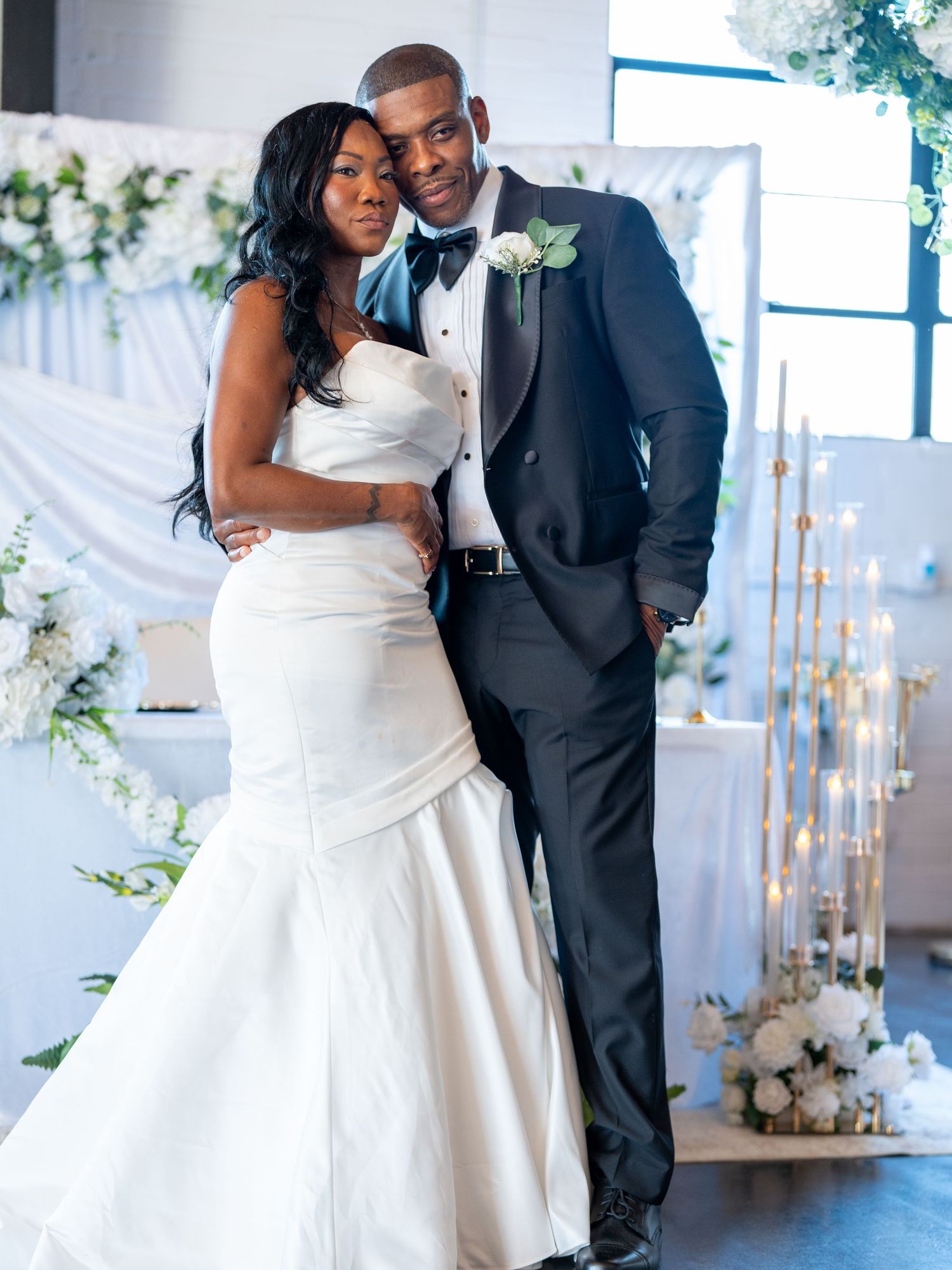 Elegant bride in white mermaid gown and groom in black tuxedo posing together at a white floral wedding reception.