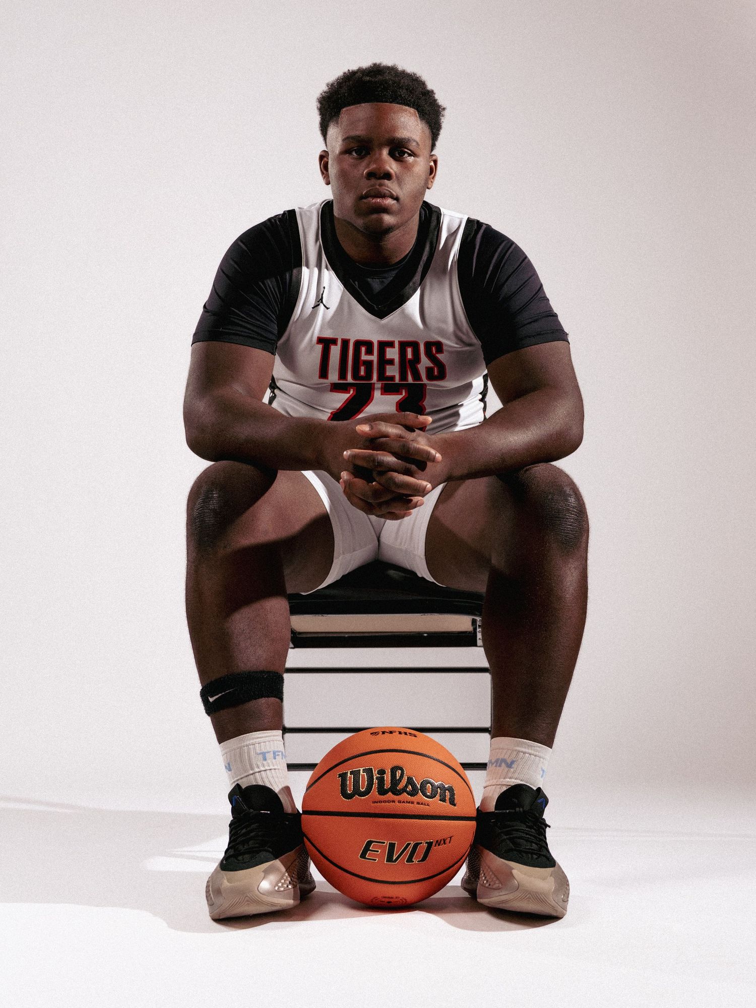 Basketball player sits in contemplative pose with Wilson basketball in studio setting against white backdrop.