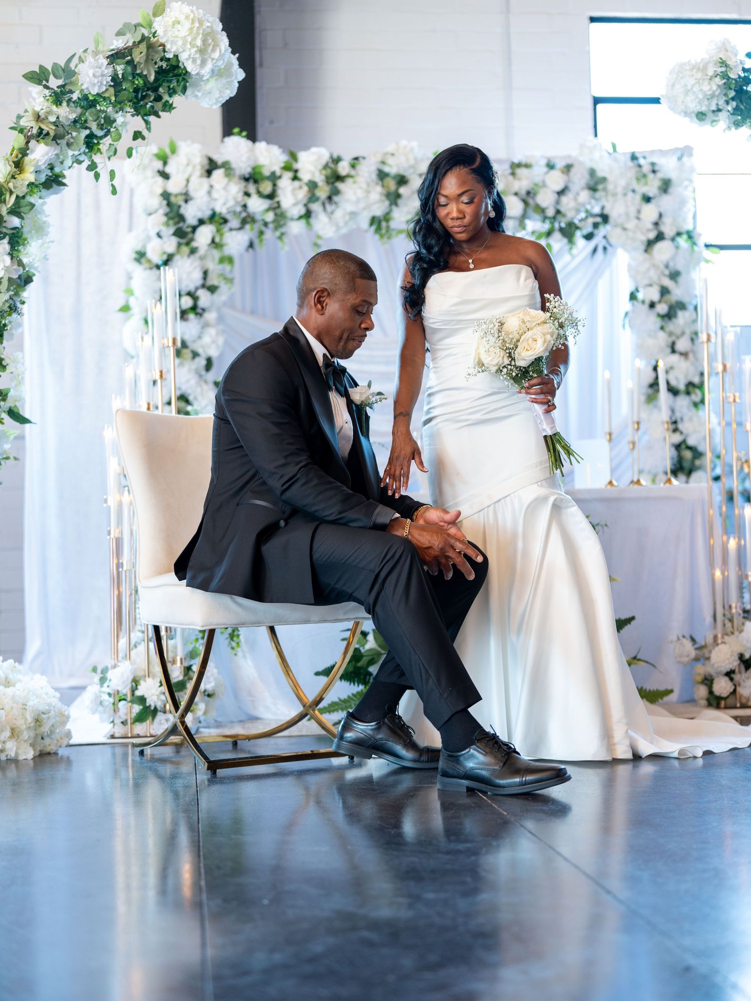 Elegant wedding couple in white gown and black tuxedo share intimate moment amid white floral arch decor.