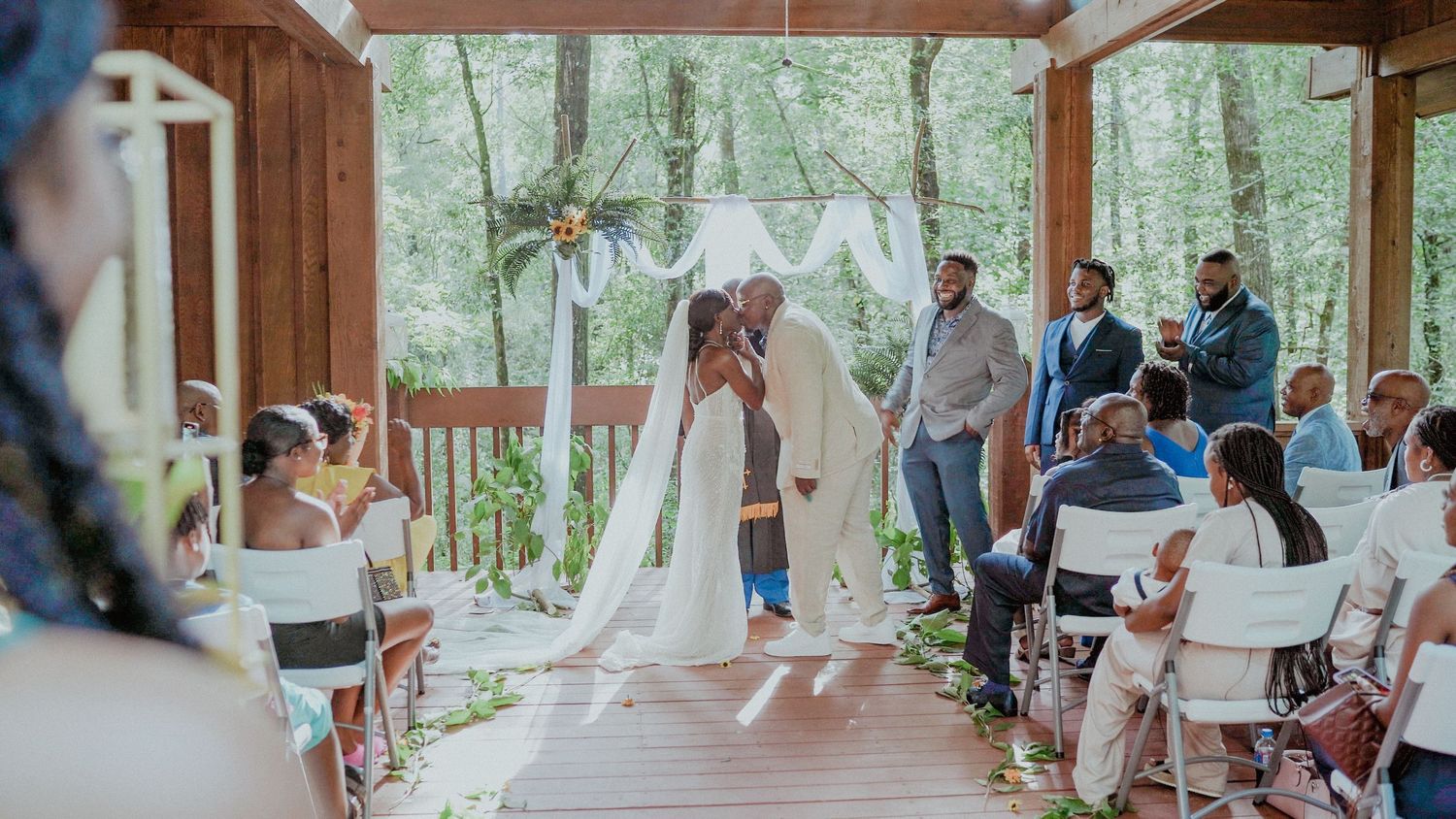 An intimate outdoor wedding ceremony taking place at a rustic wooden pavilion in the forest with guests seated on white chairs.