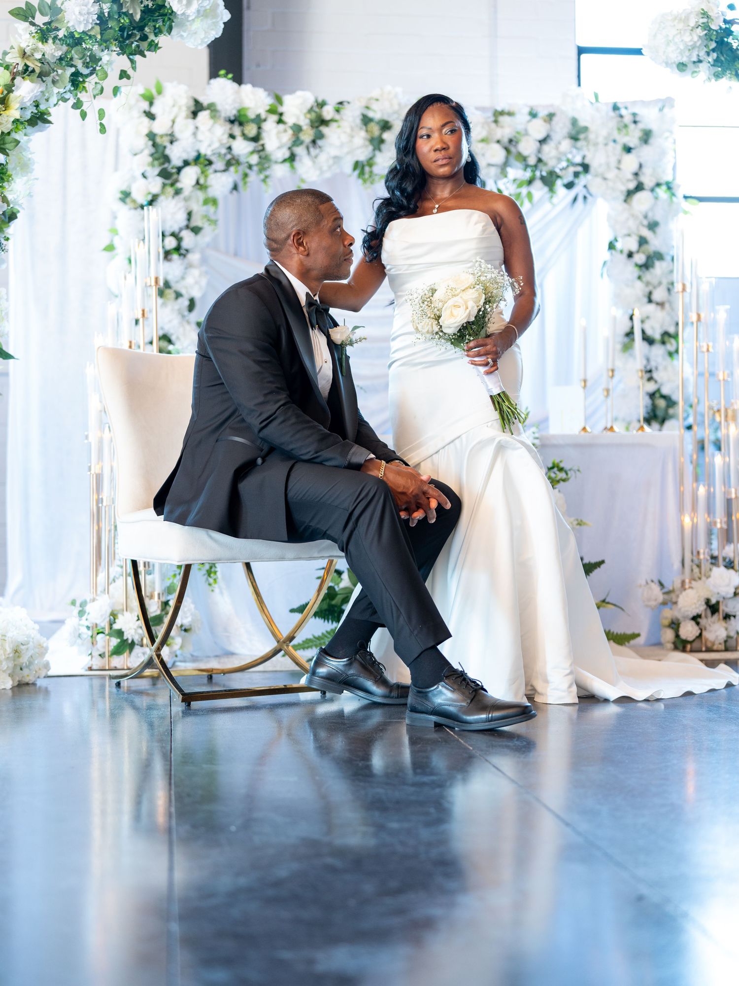 Elegant wedding couple posing in white floral venue, bride in strapless gown holding bouquet, groom in black tuxedo.