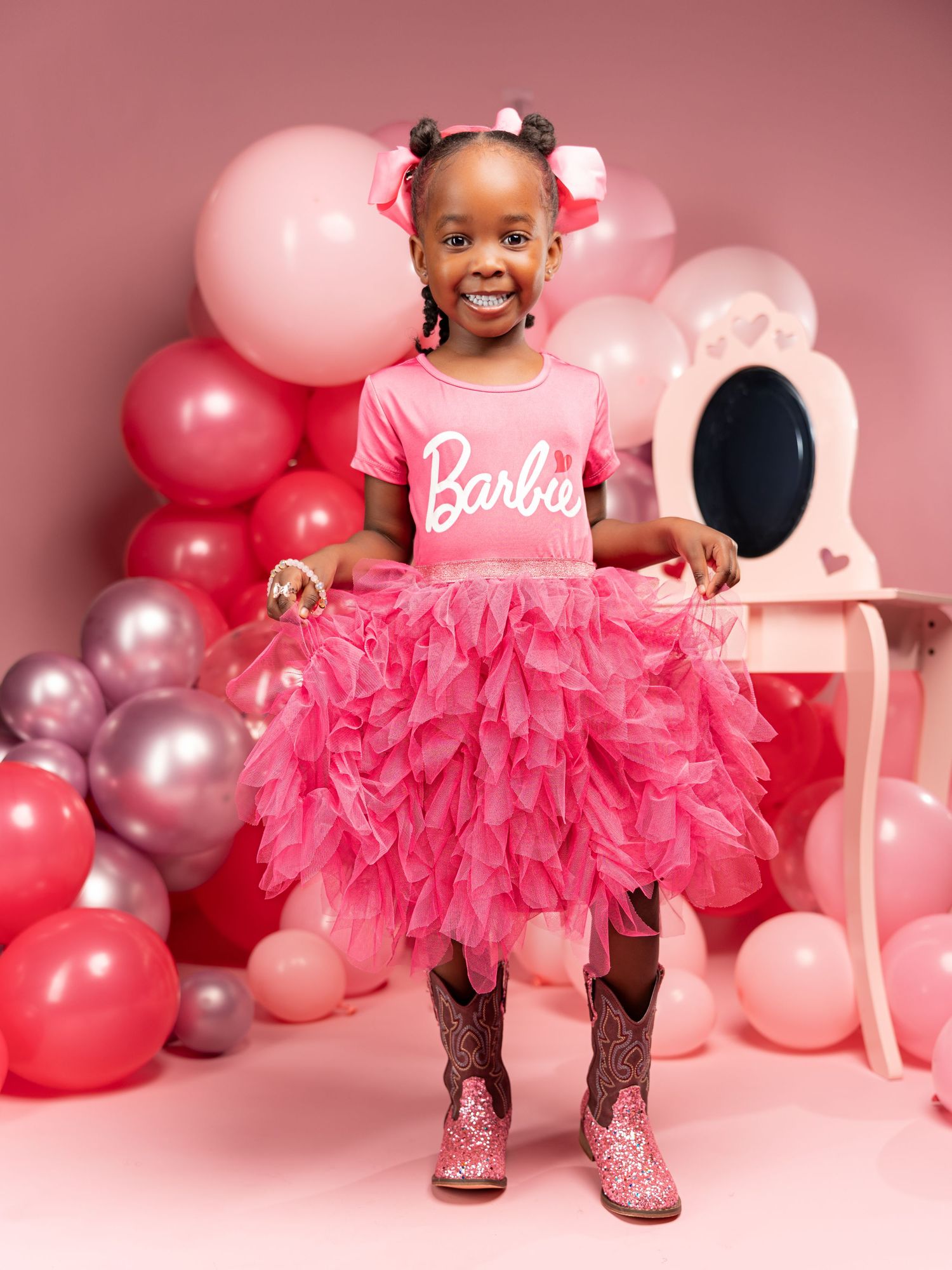 A child in a pink tulle dress and sparkly boots poses against a pink backdrop with silver and pink balloons.
