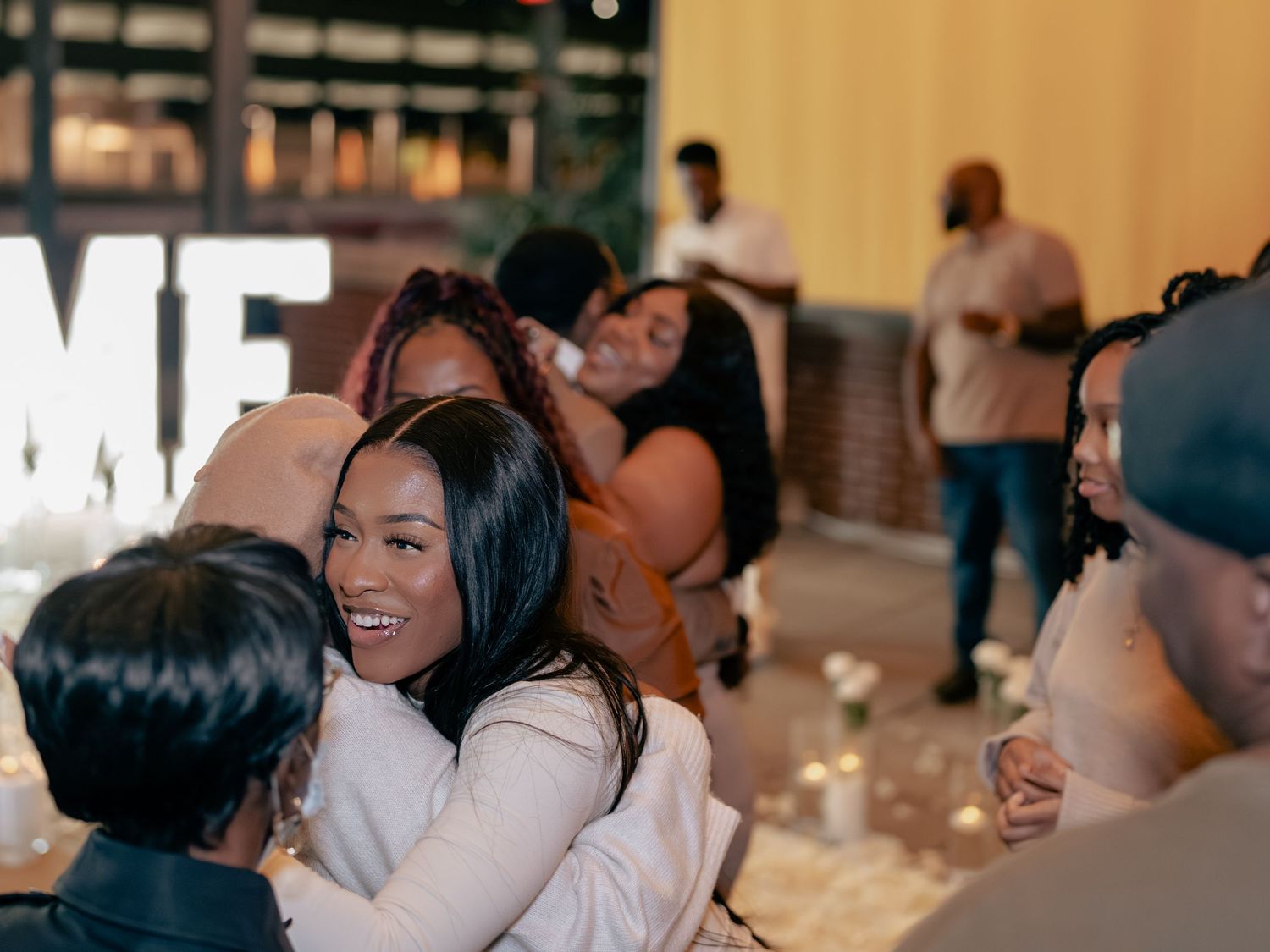 People smiling and hugging at a warm indoor gathering, with decorative lights and guests socializing in the background.