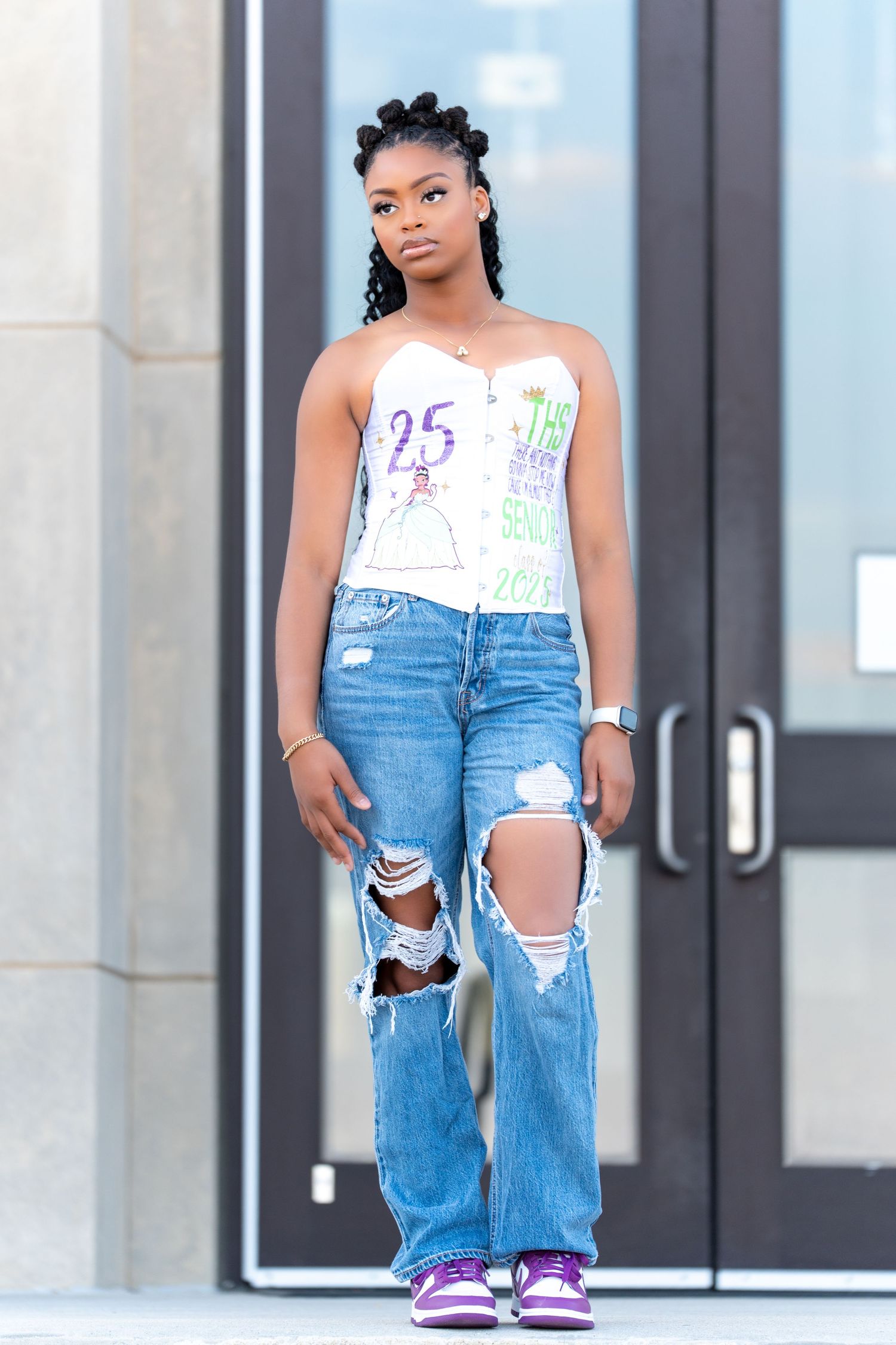 Woman in white tank top and ripped blue jeans poses confidently outside a building entrance.