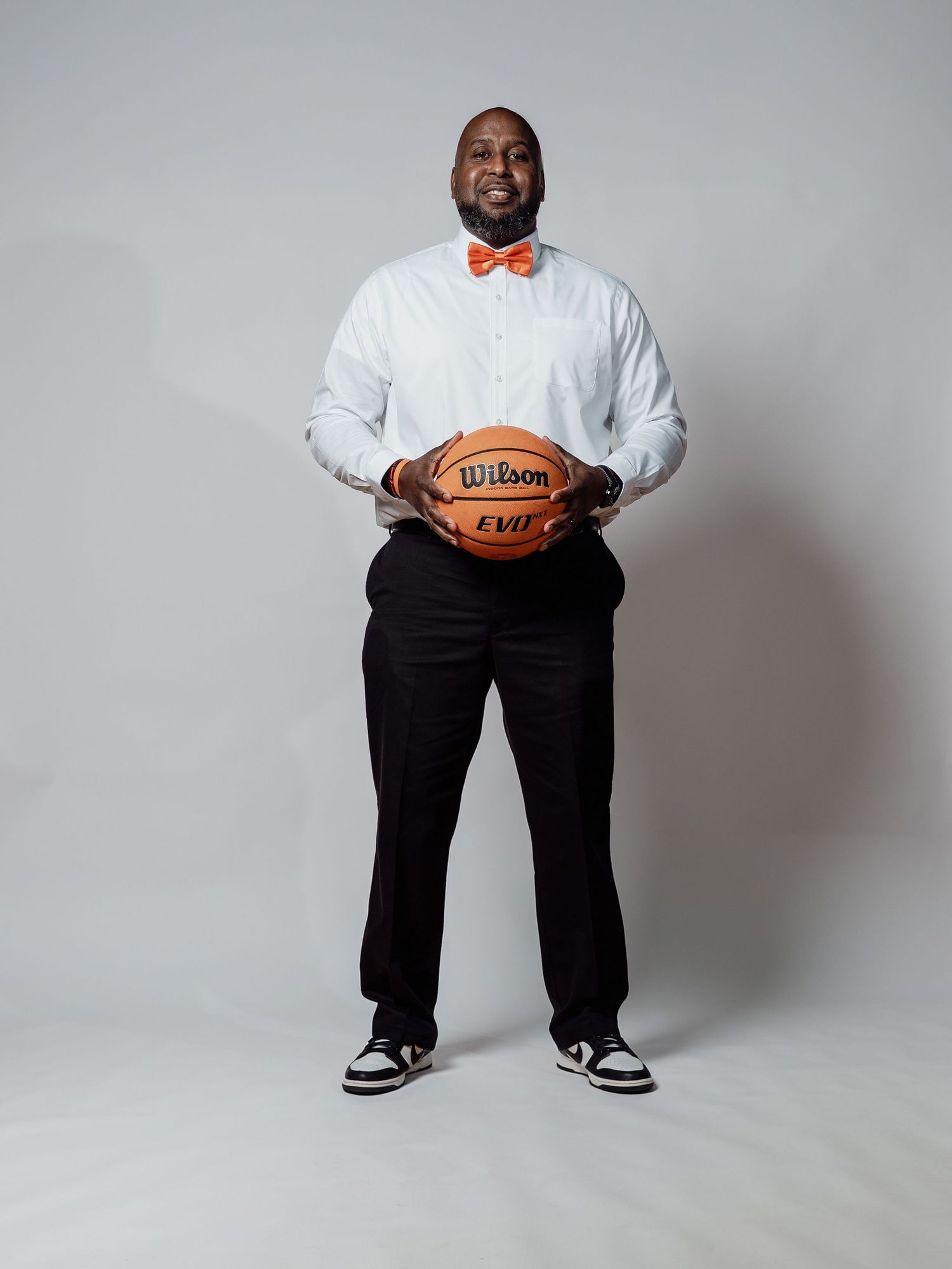 Man in a white dress shirt, orange bow tie, black pants and sneakers stands smiling against a gray backdrop holding a Wilson basketball.