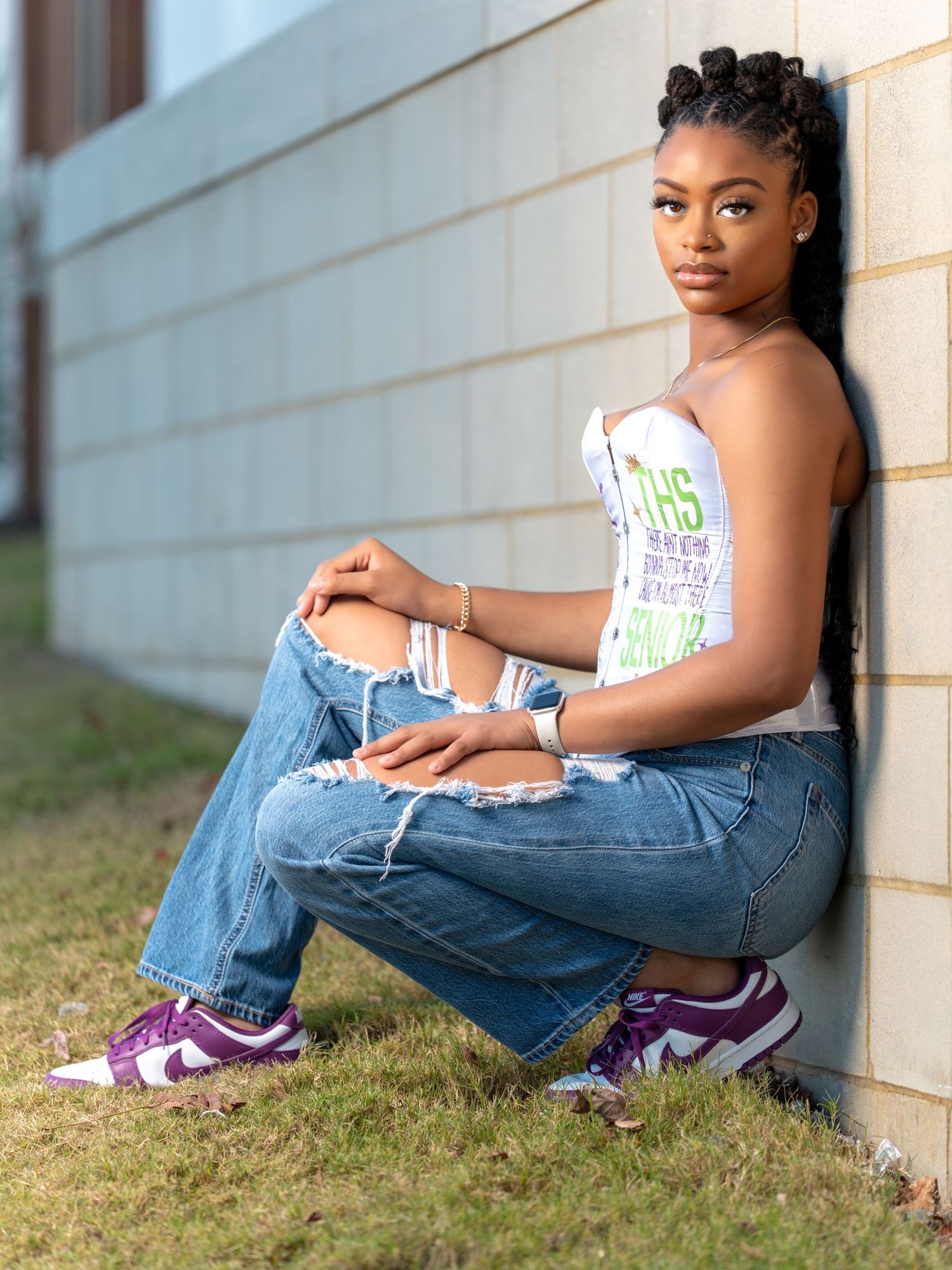 A person in a white tank top and distressed jeans poses casually against a wall while wearing purple sneakers.