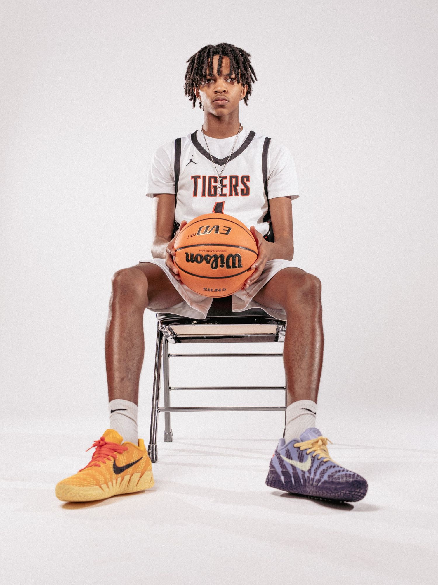 Player sits on metal chair holding basketball while wearing Tigers uniform and colorful sneakers.