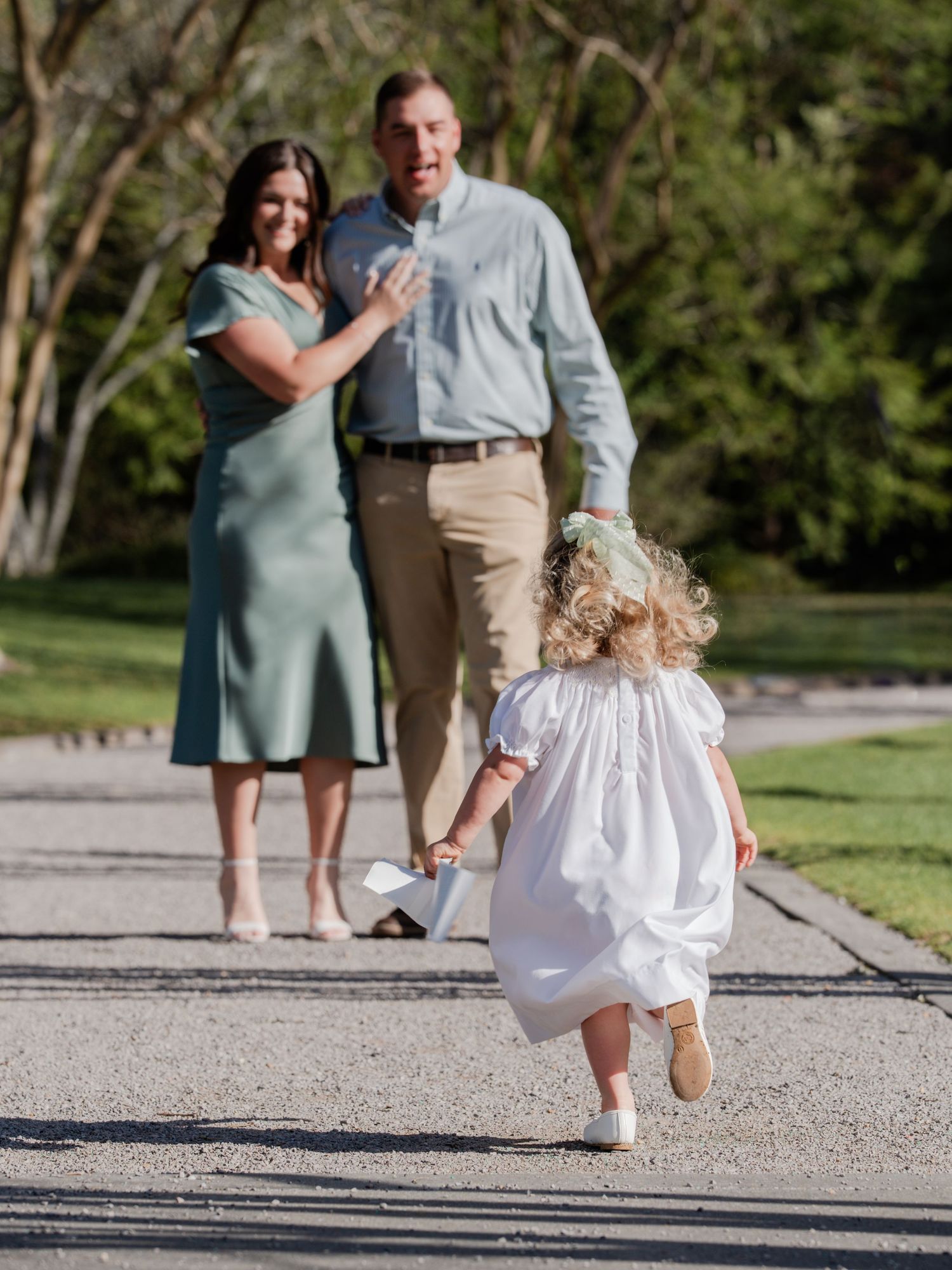 A toddler in a white dress runs along a garden path toward a smiling woman in a green dress and a laughing man in khakis.