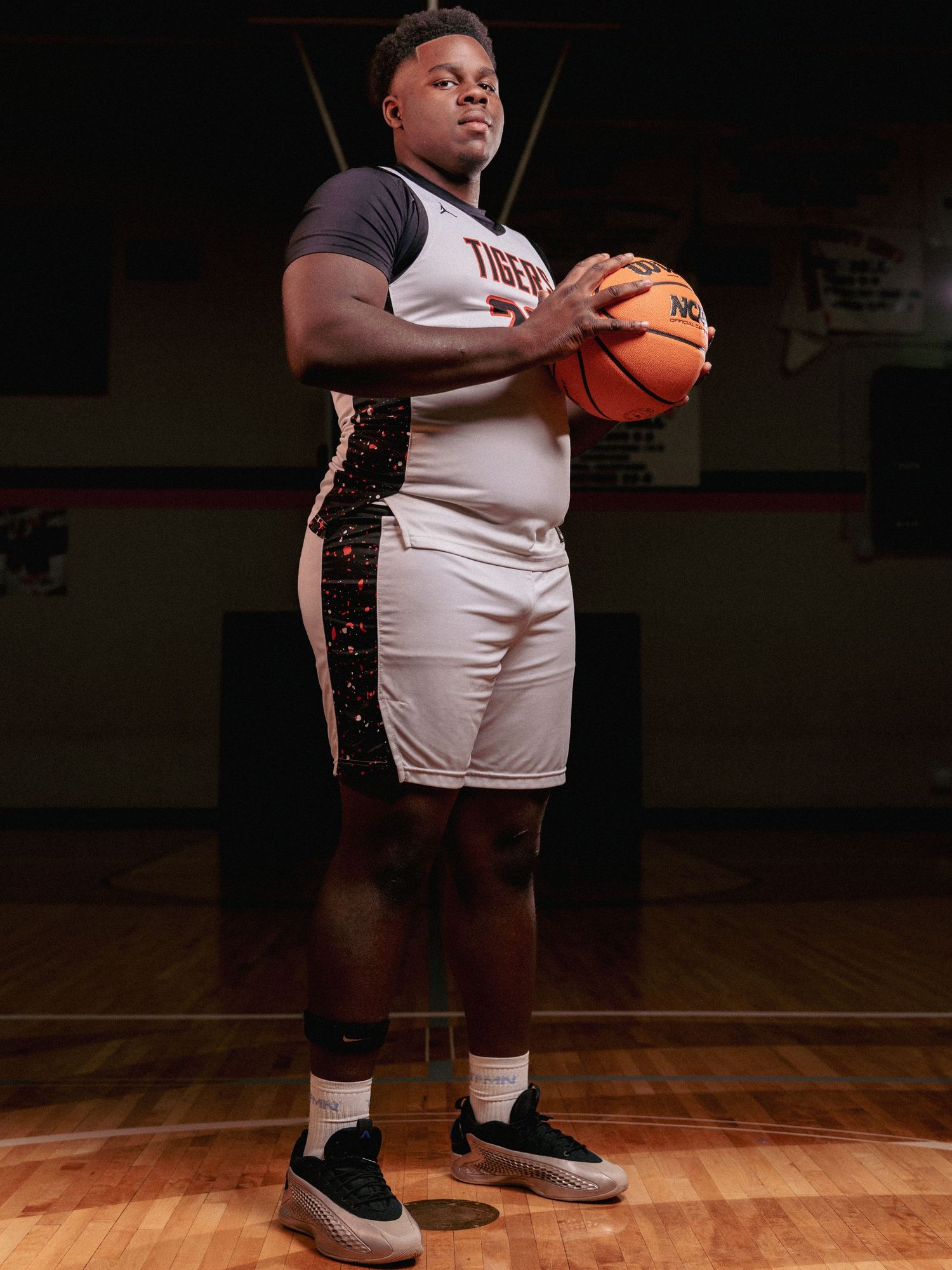 Basketball player in white uniform stands on wooden court holding ball in dramatic lighting.
